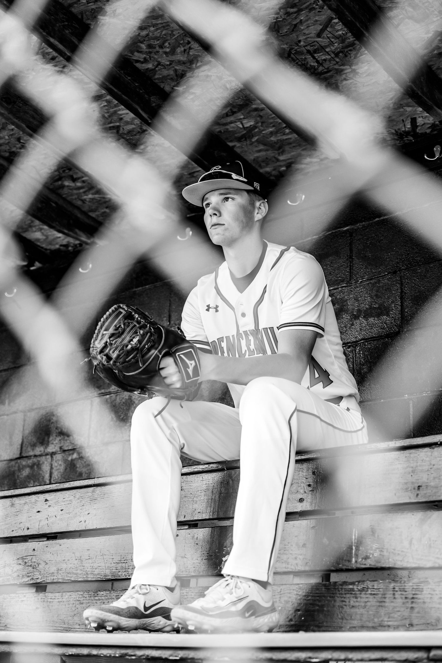 Baseball players in white uniforms sit in the dugout behind protective netting during a game.