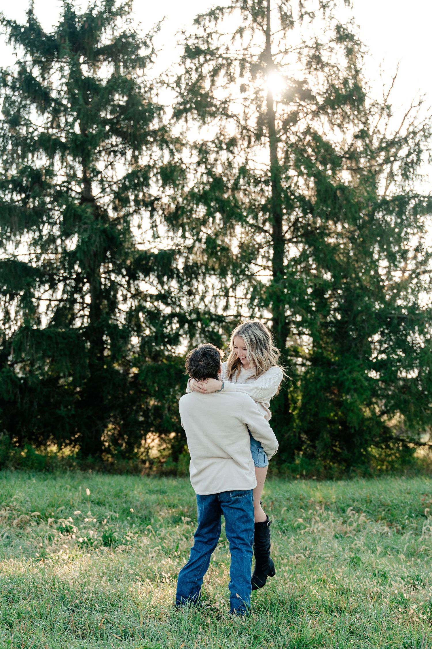 Two people embrace in a grassy field at sunset with evergreen trees silhouetted in the background.