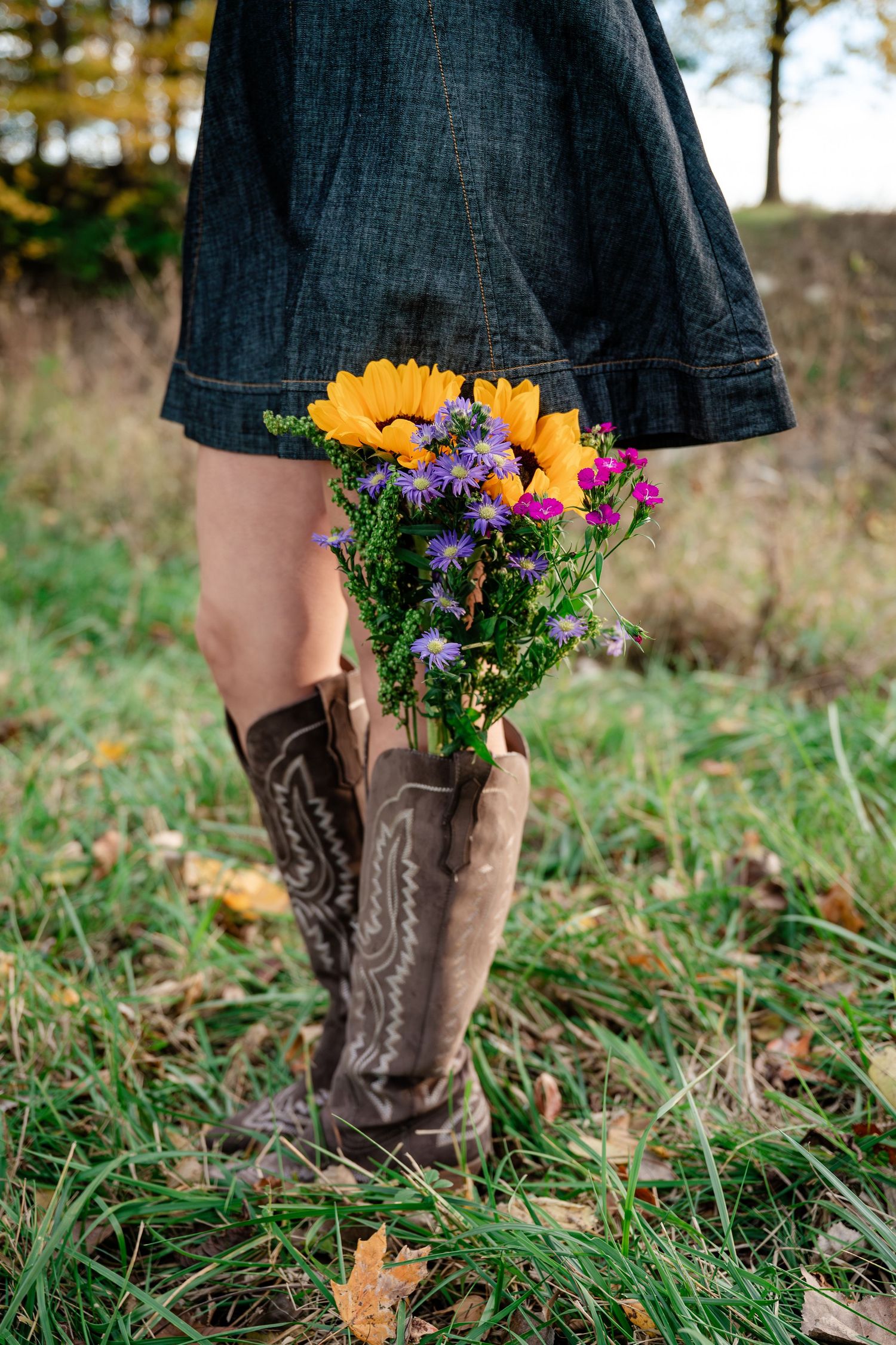 Brown cowboy boots with a black dress and wildflower bouquet stand in green grass during autumn.