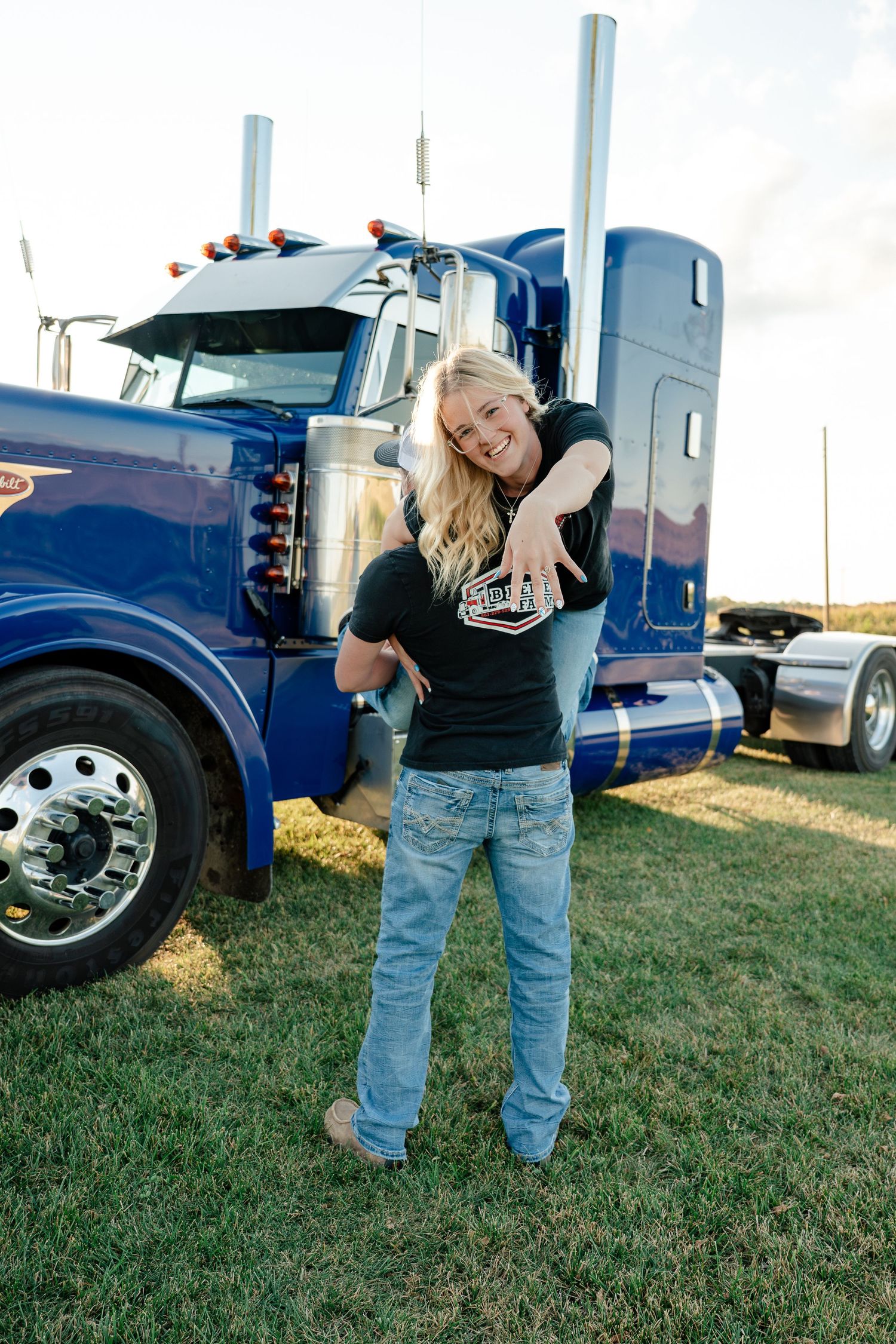 A person in jeans and a black shirt poses near blue semi trucks parked on grass at a truck stop.