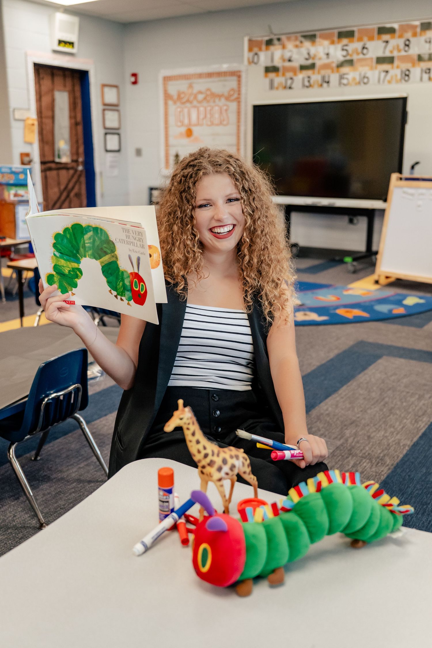 A teacher demonstrates The Very Hungry Caterpillar story with a plush toy in a classroom setting.
