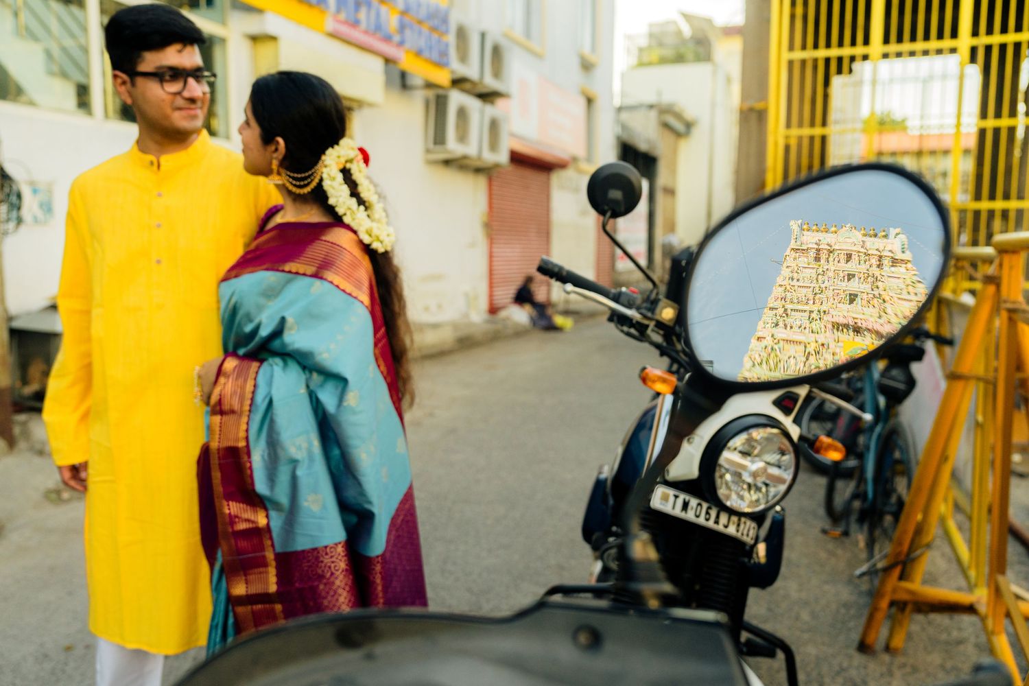 Indian couple in traditional attire near a motorcycle, temple reflected in mirror, colorful cultural clothing.