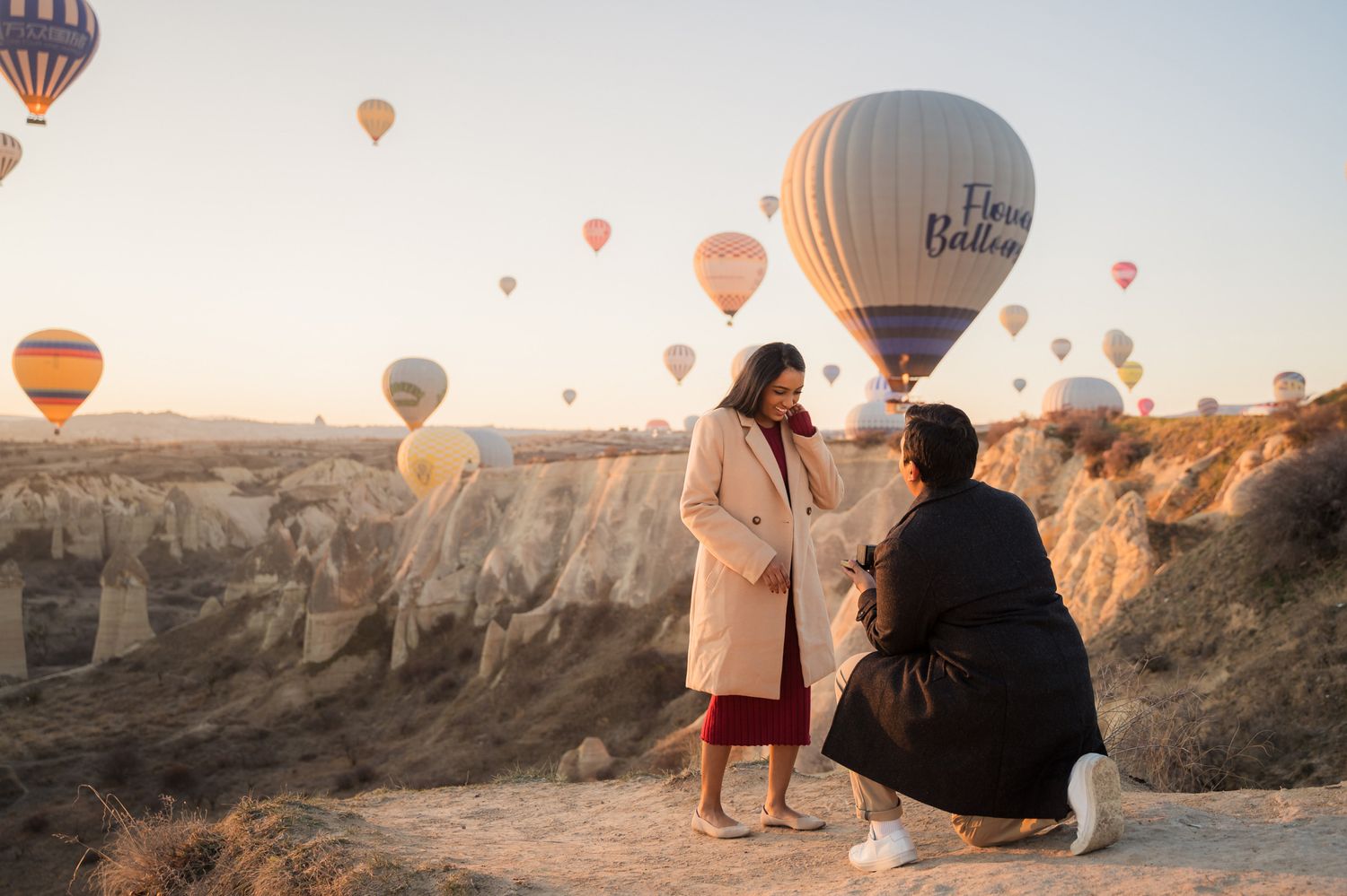 Hot air balloons float over the rocky landscape of Cappadocia at sunrise while tourists watch from a viewpoint.