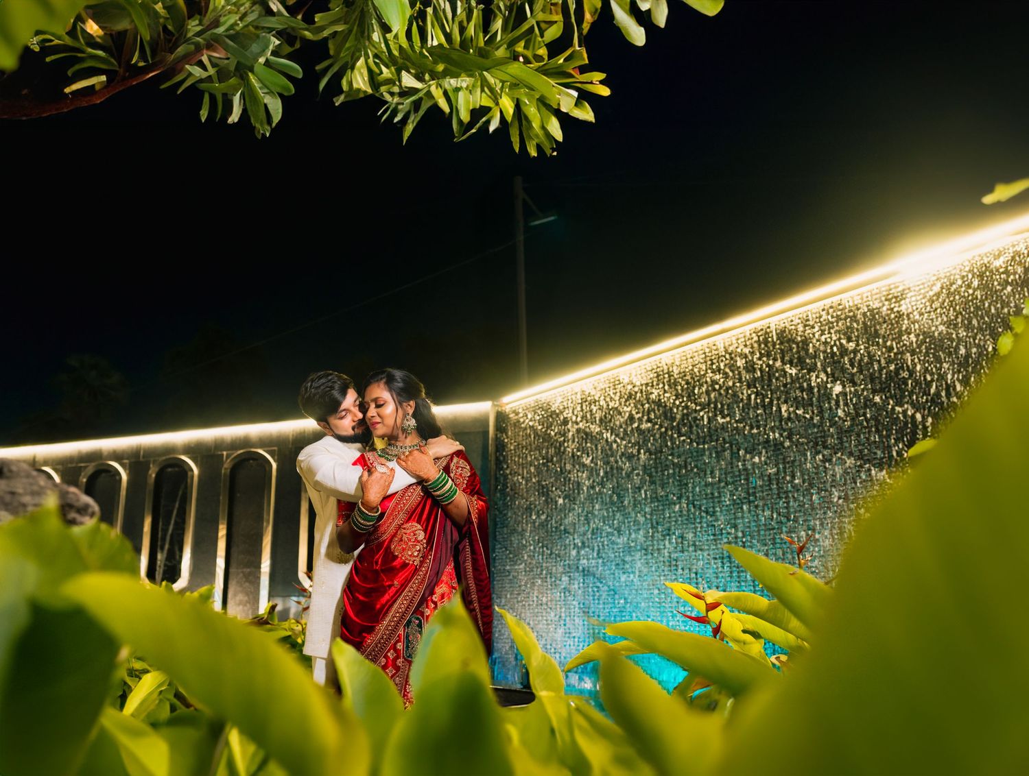 Couple poses near illuminated bridge railing at night through leafy branches.