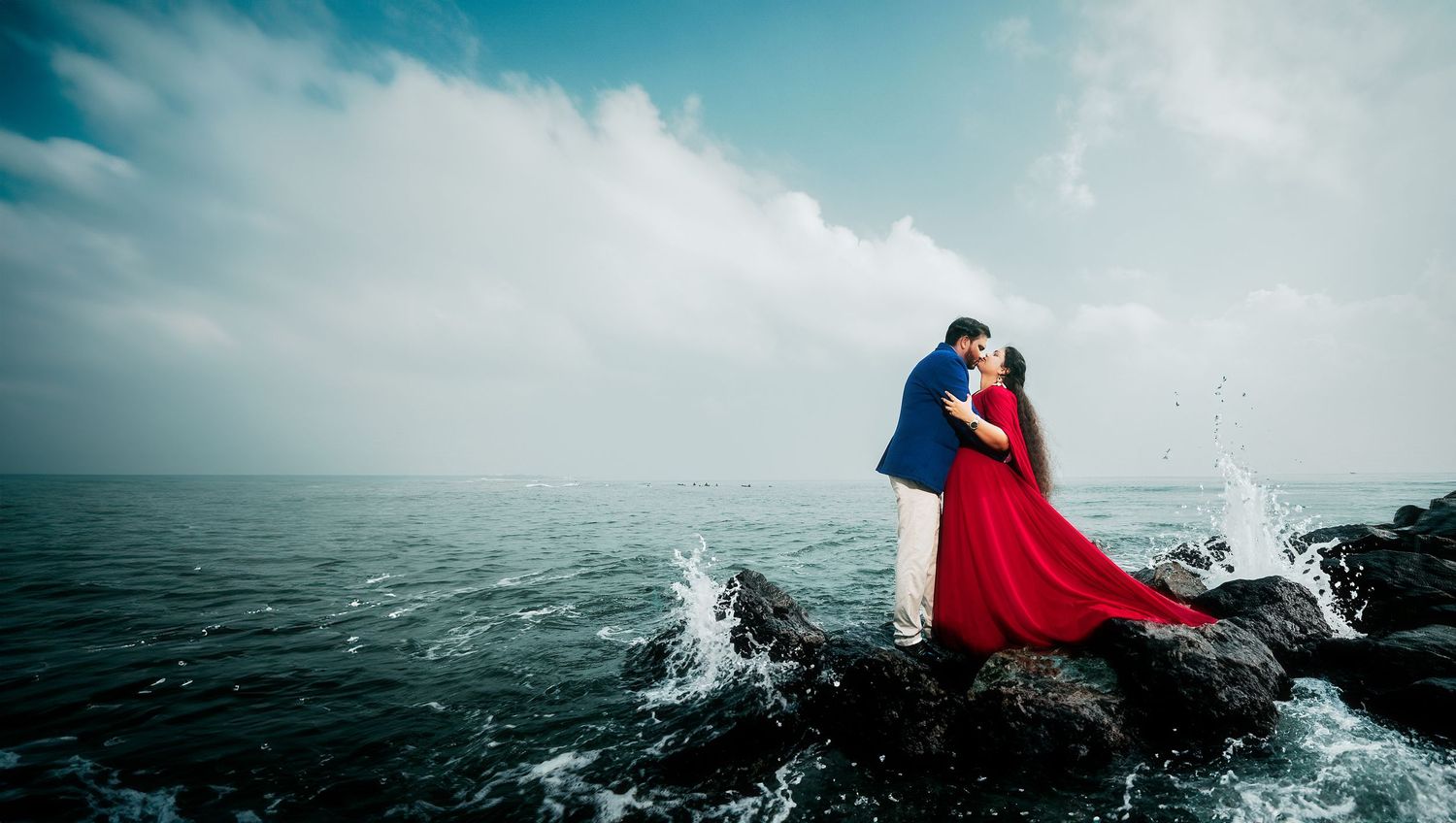 A dramatic coastal photoshoot featuring a couple in blue and red attire standing on rocks with crashing waves.