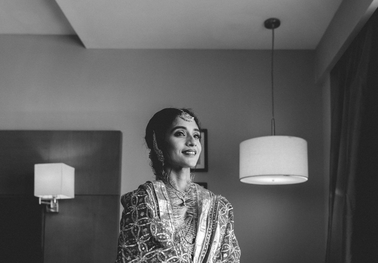Black and white photo of someone in traditional South Asian attire smiling with pendant lights visible in the background.