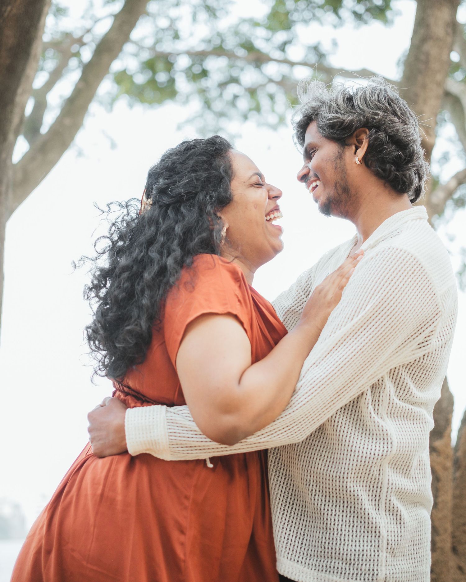 A couple embraces lovingly outdoors among trees with natural sunlight filtering through.