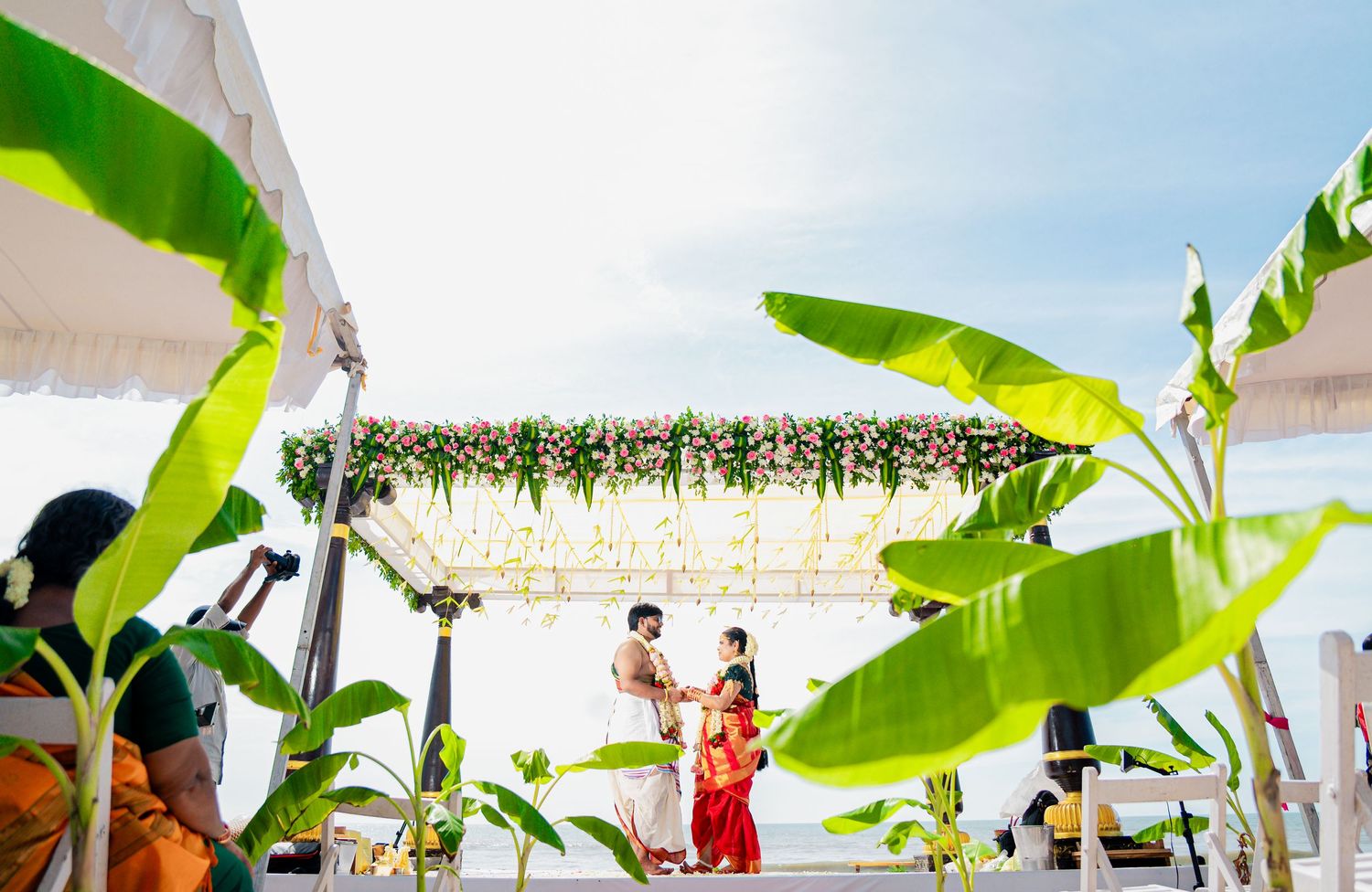 Tropical wedding ceremony setup with banana leaves and floral arrangements under white canopy.