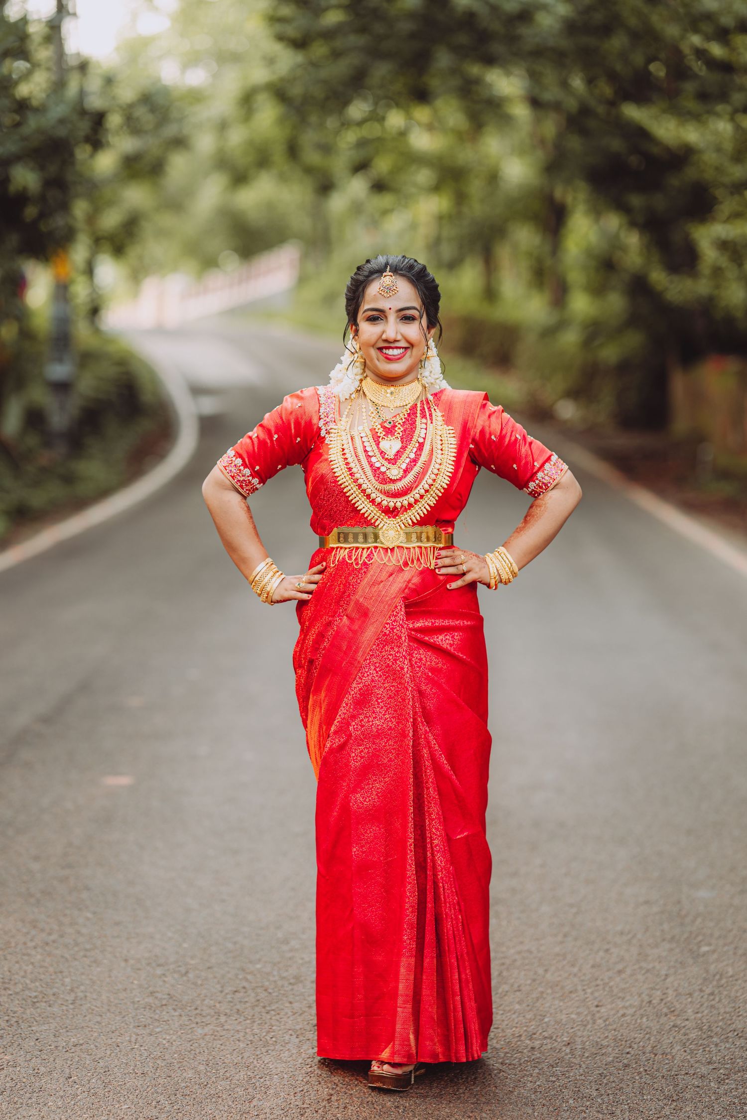 Traditional South Indian bride in bright red saree with gold jewelry poses on a scenic road.
