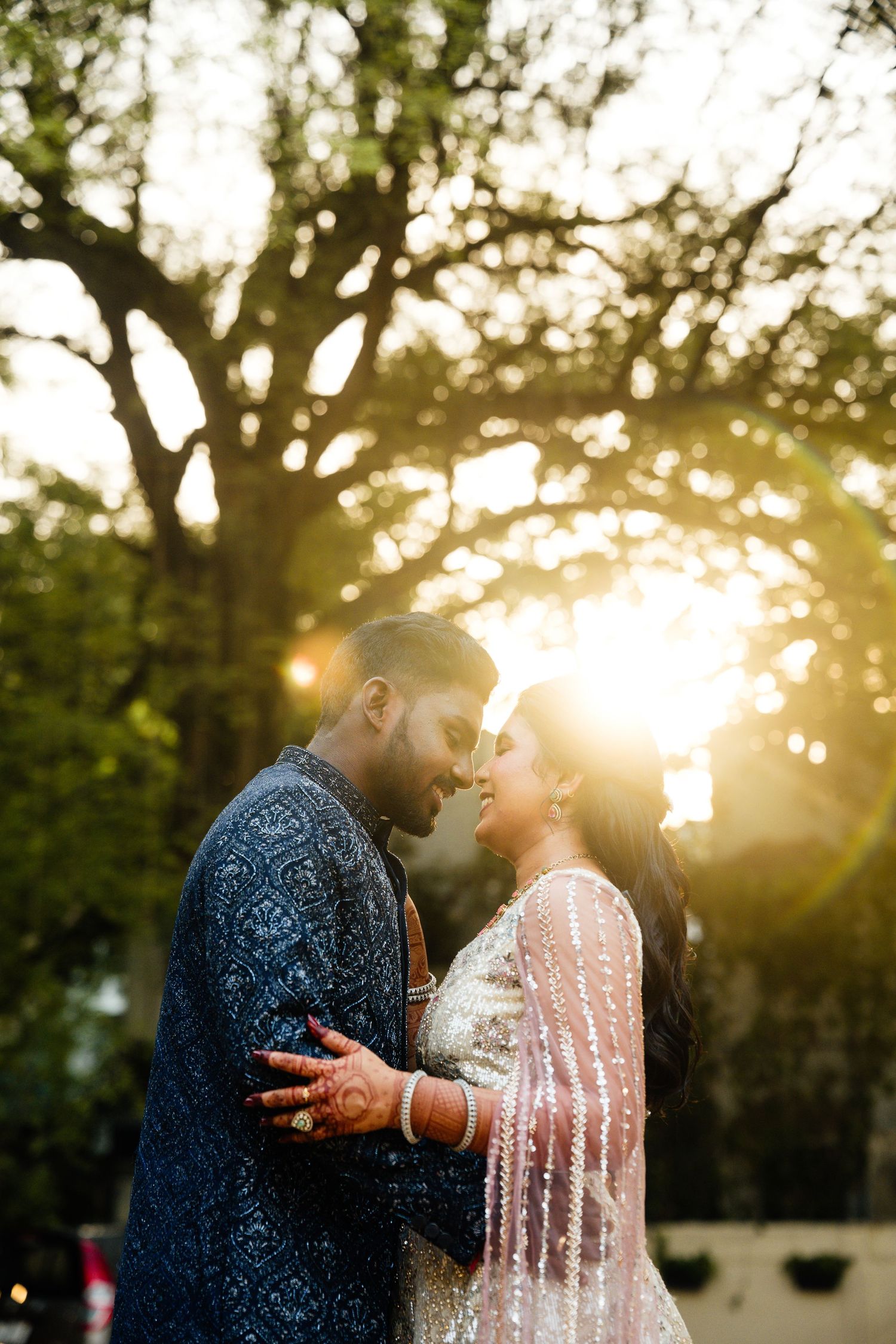Couple in traditional South Asian wedding attire sharing a romantic moment outdoors with golden sunset light filtering through trees.
