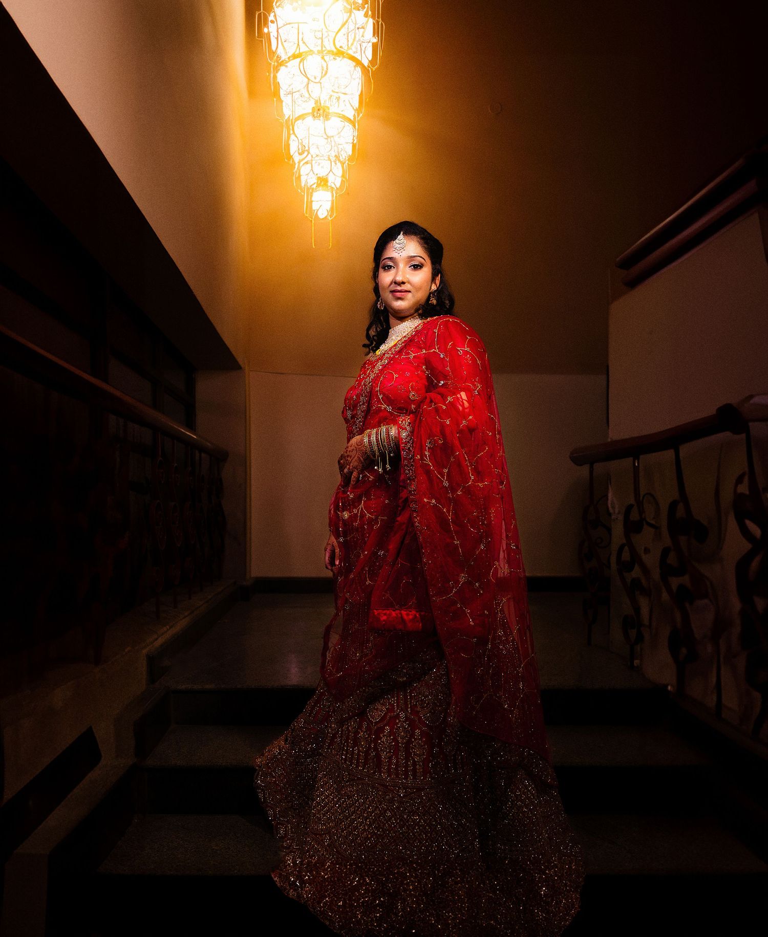 A dramatic portrait of someone wearing a red sequined lehenga and dupatta standing on stairs under warm lighting.