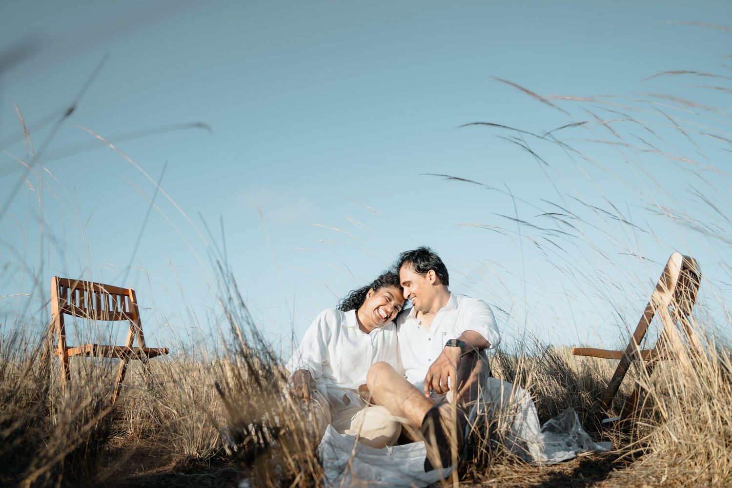 Couple sharing tender moment among tall grass with rustic wooden chair in background.