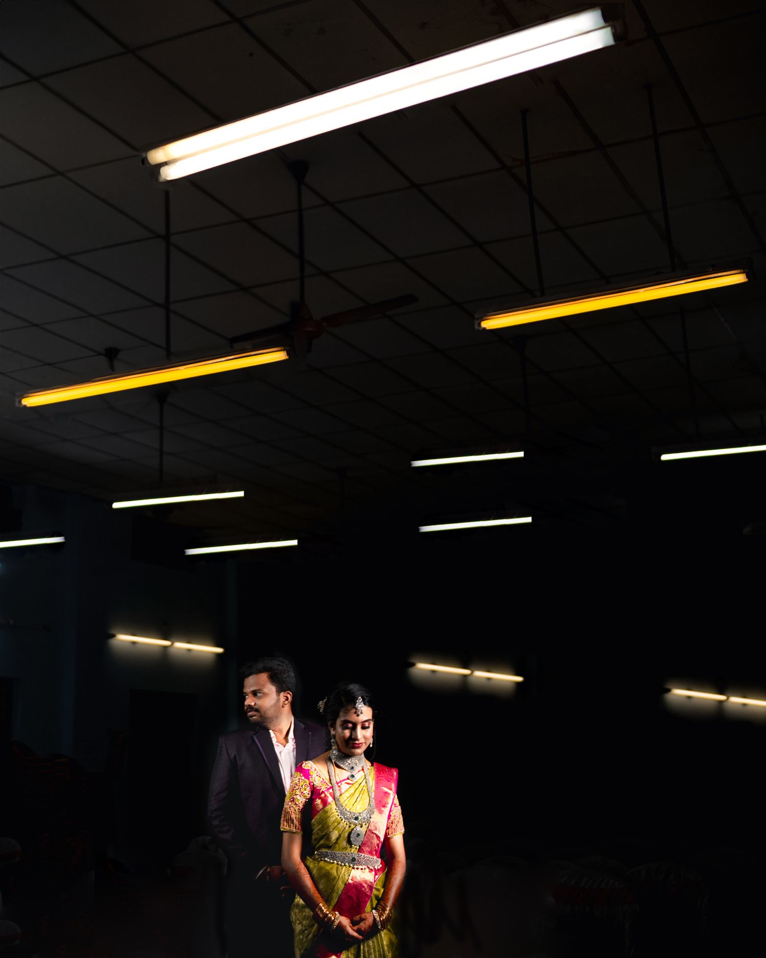 Stylish couple poses under geometric lighting pattern in dark indoor space.