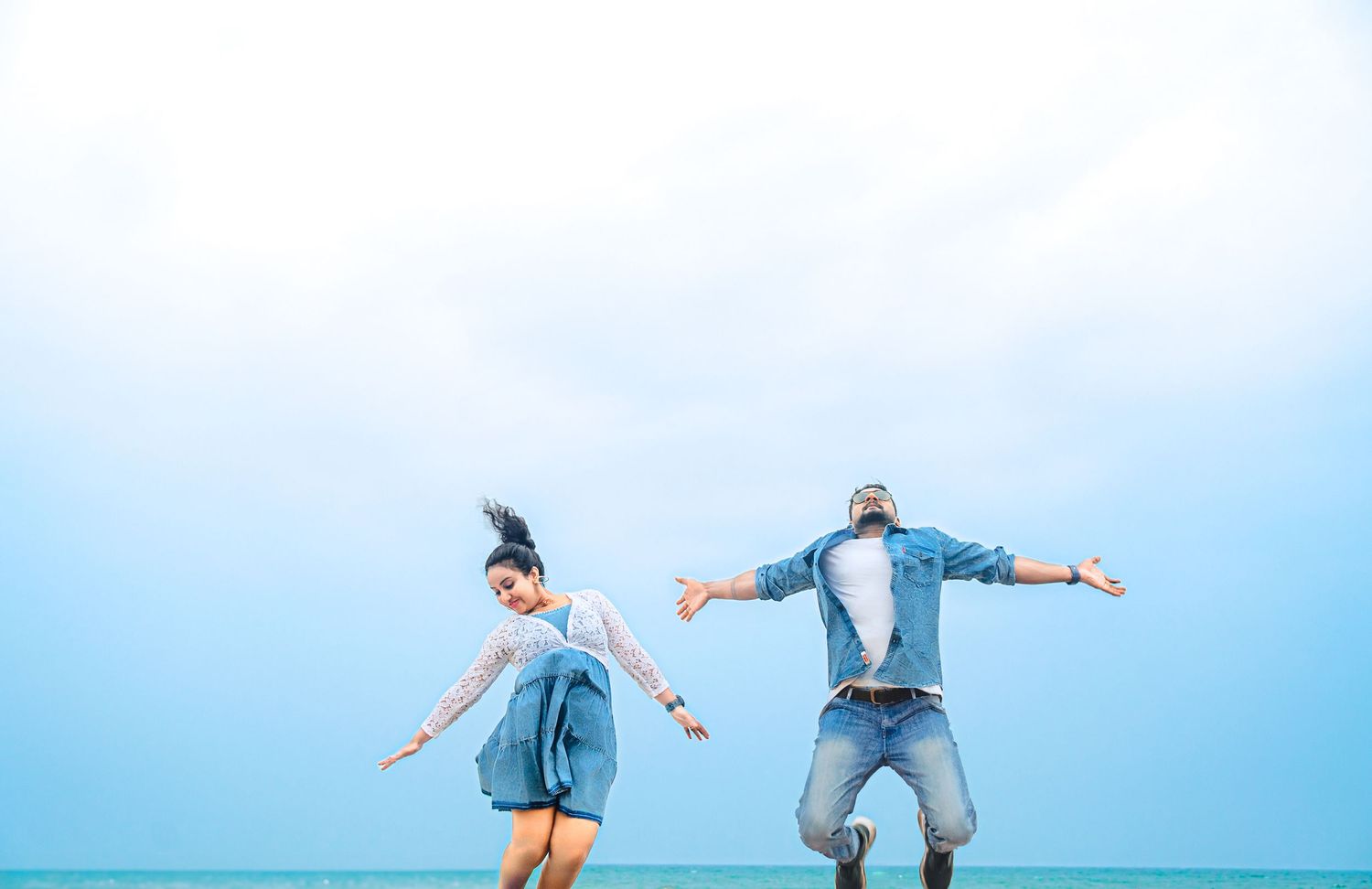 Couple jumping with arms spread against blue sky backdrop.