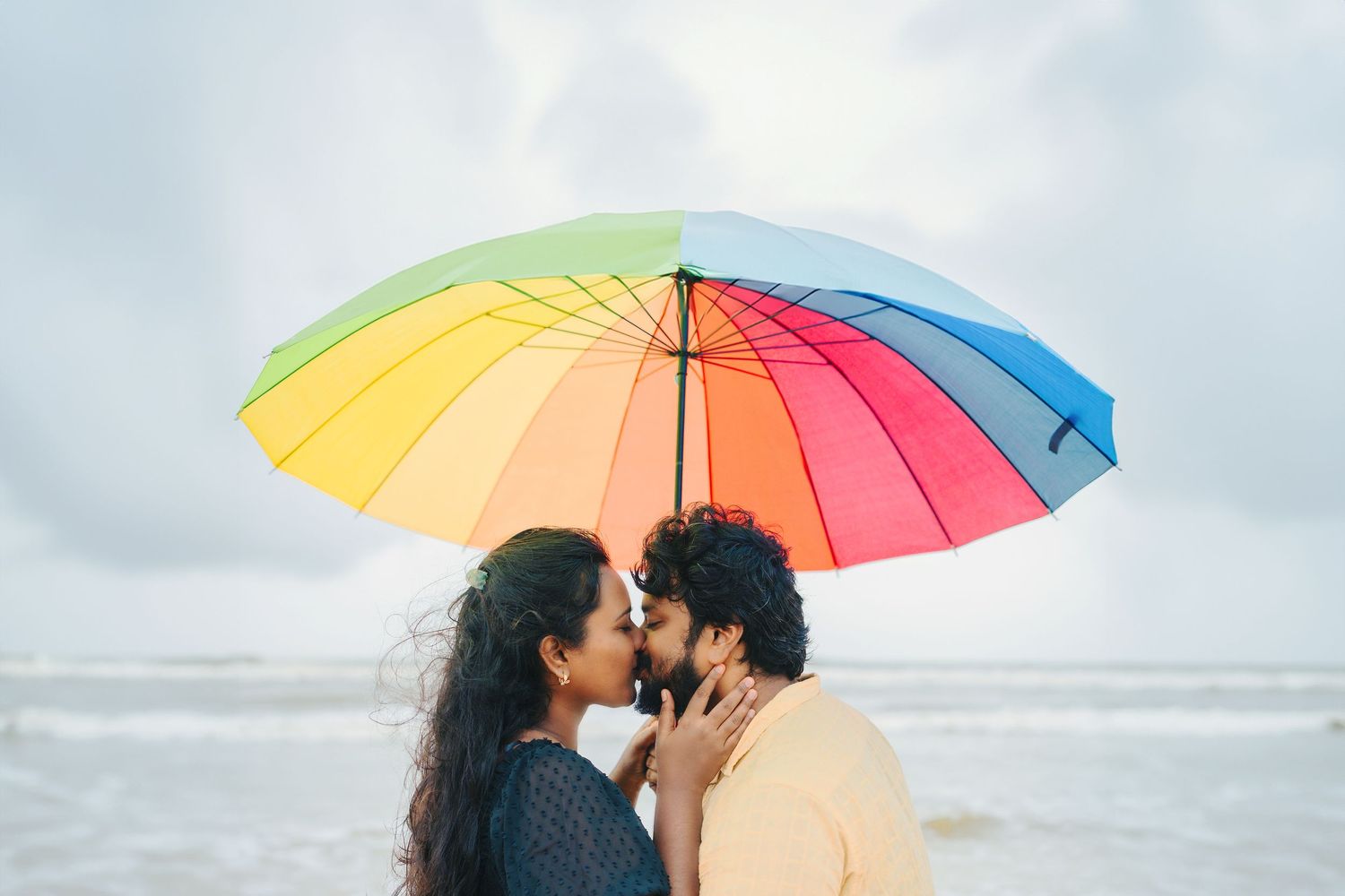 A romantic couple shares an intimate moment under a colorful rainbow umbrella at the beach.