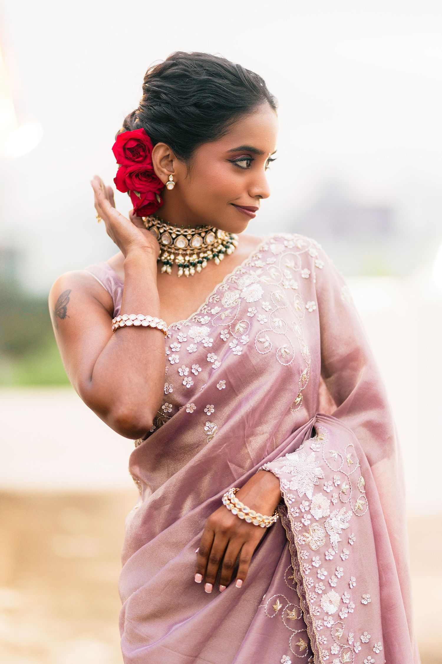 Woman in pink embroidered saree with traditional Indian jewelry poses elegantly against light background.