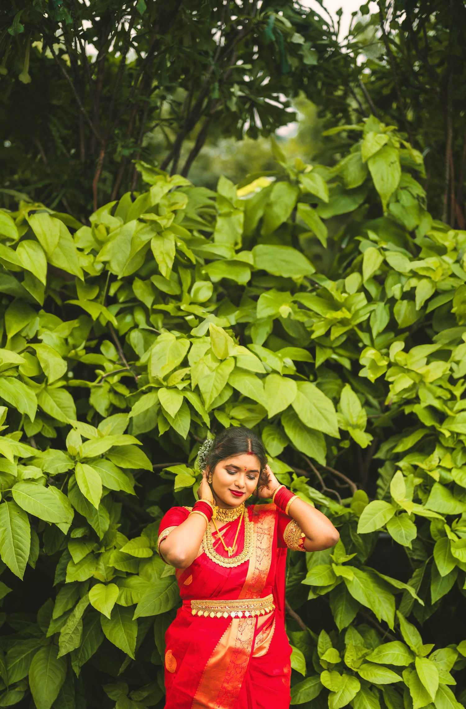 Traditional red saree-clad figure posing against lush green foliage background.