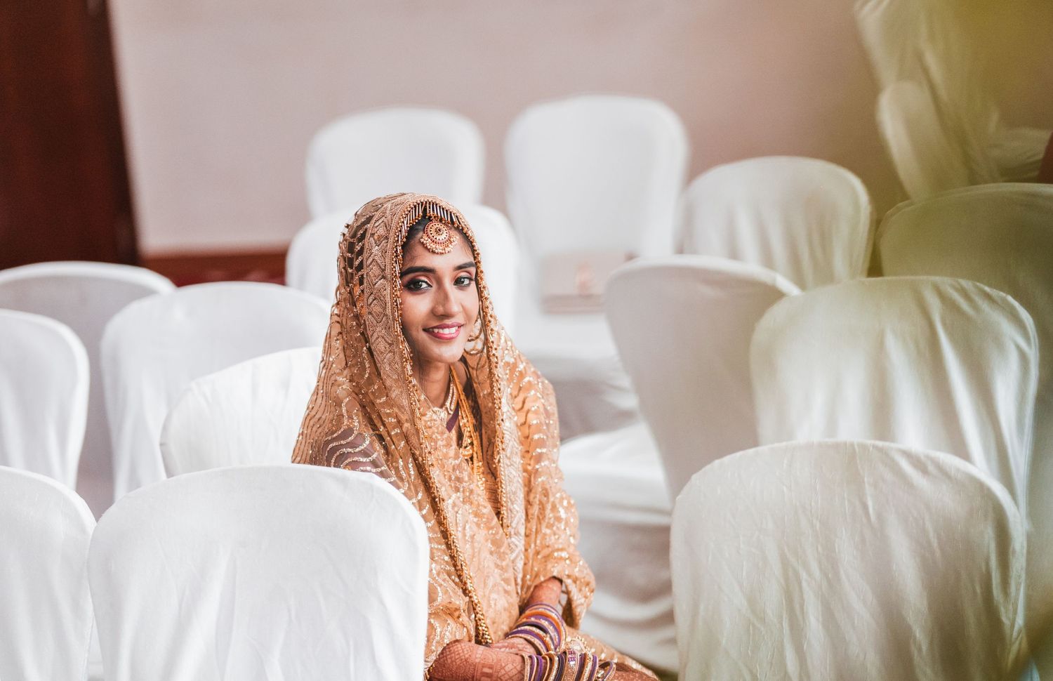 Person wearing a peach-colored traditional veil sits among white chairs in an indoor setting.