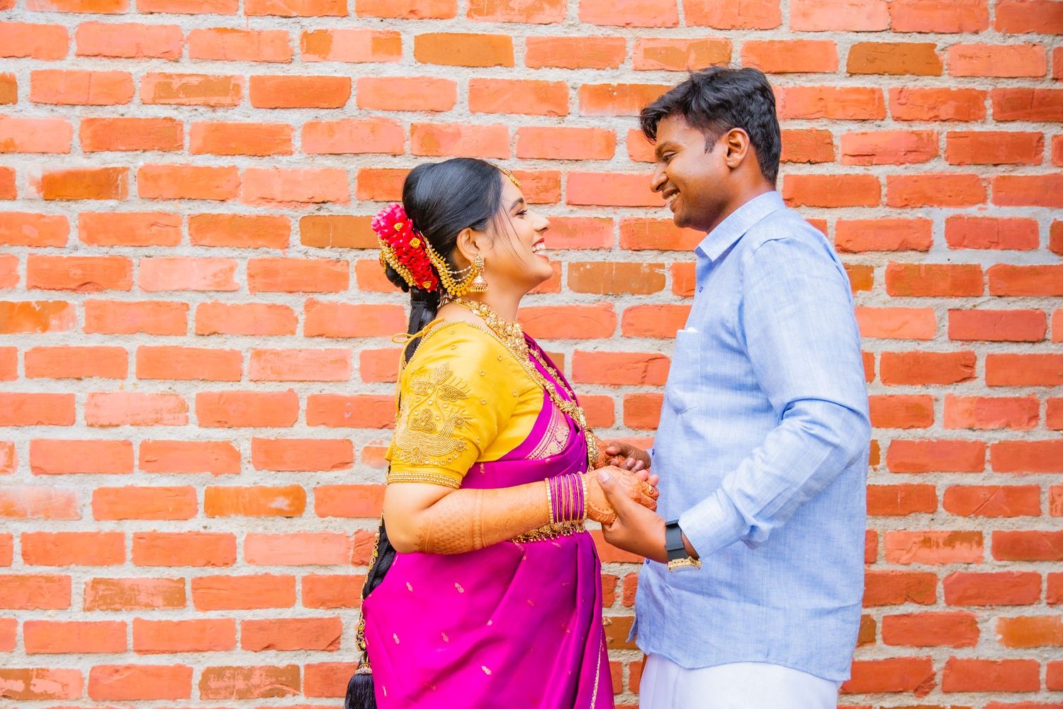 A couple shares an intimate moment against a red brick wall wearing traditional South Indian clothing.