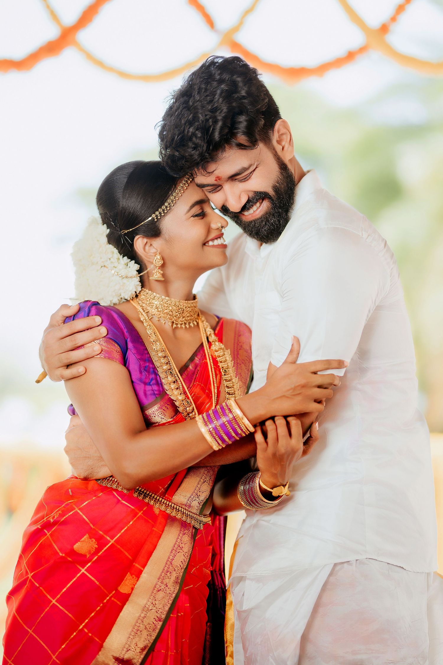 Traditional South Indian wedding couple in festive attire embracing under decorative backdrop.