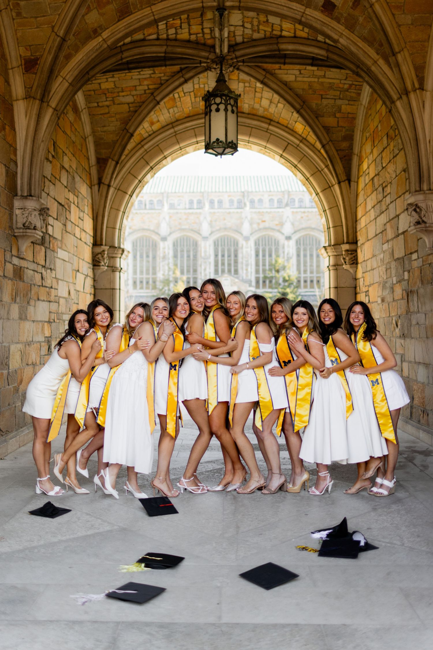 A group in white dresses and yellow stoles poses together under a stone archway in a graduation photo series.