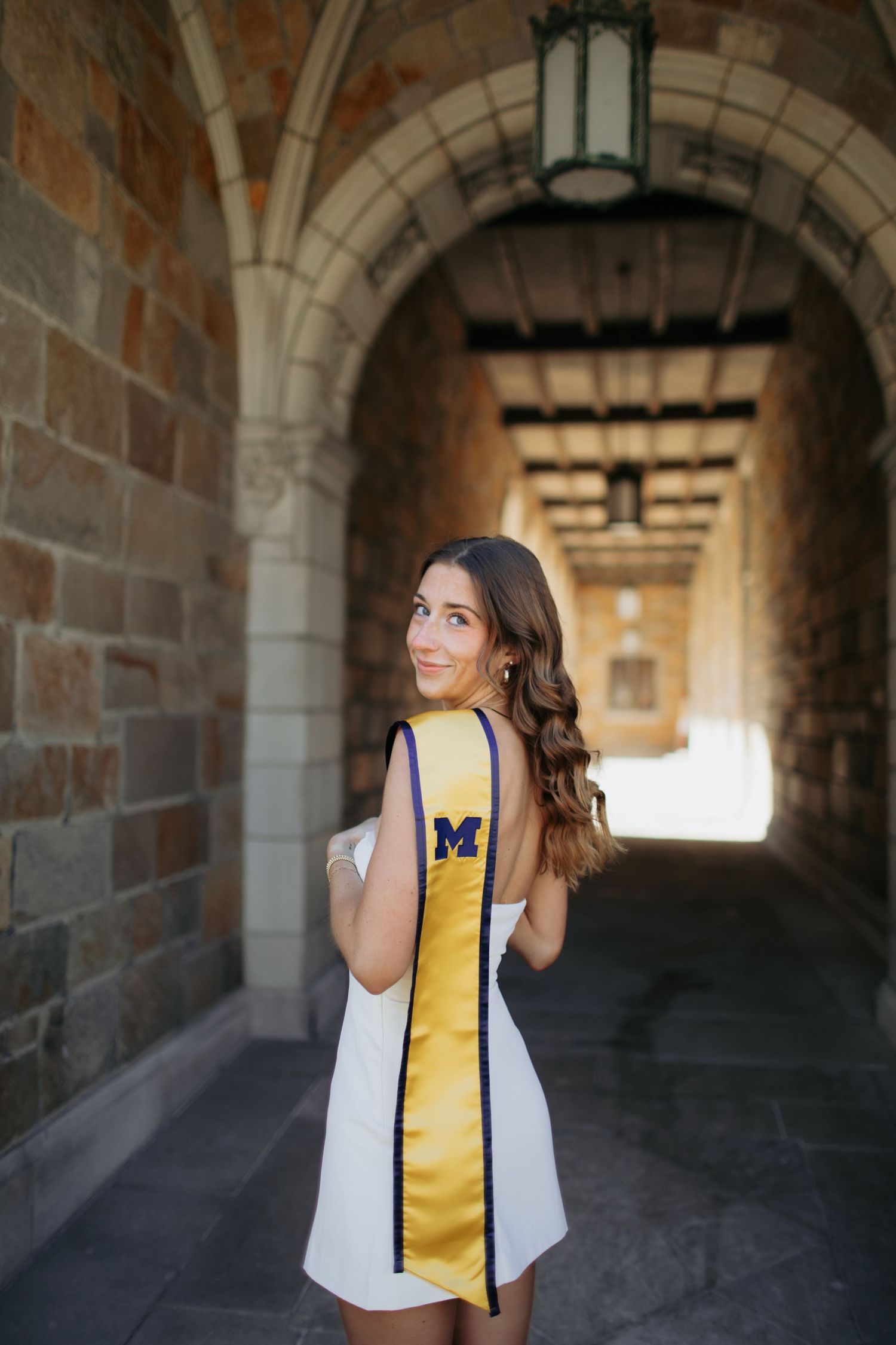 Graduate wearing white dress and yellow University of Michigan stole poses in stone archway hallway.