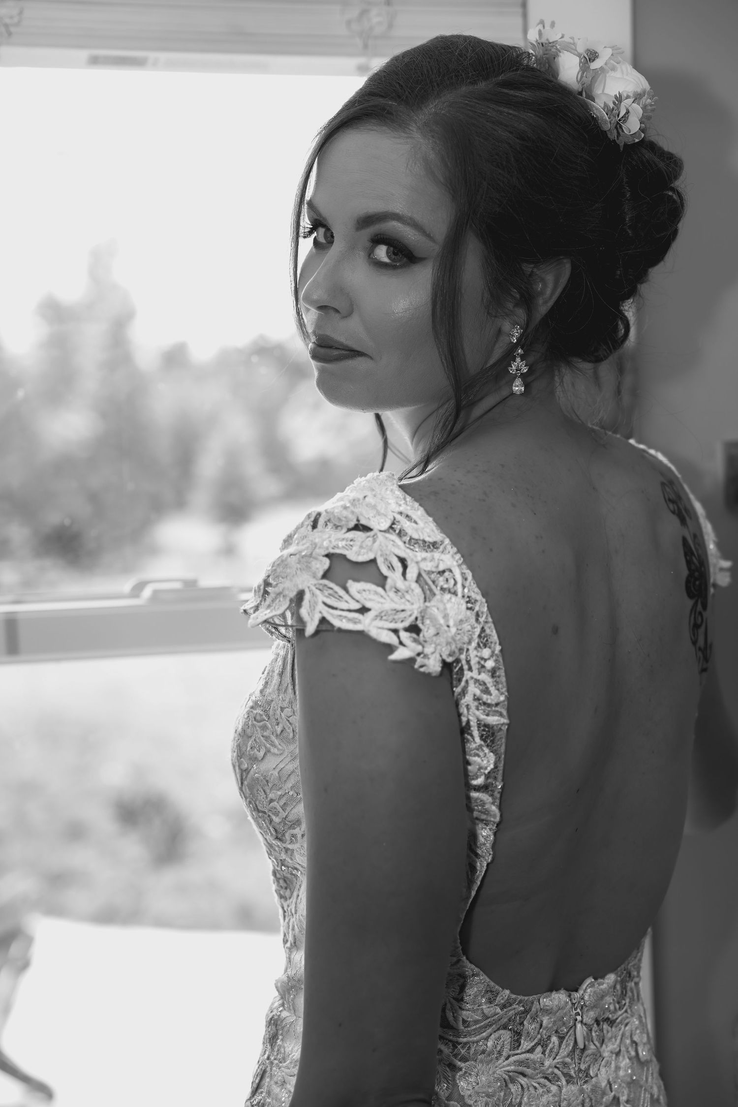 Black and white photo of a bride in a backless lace wedding gown, looking over her shoulder near a window.