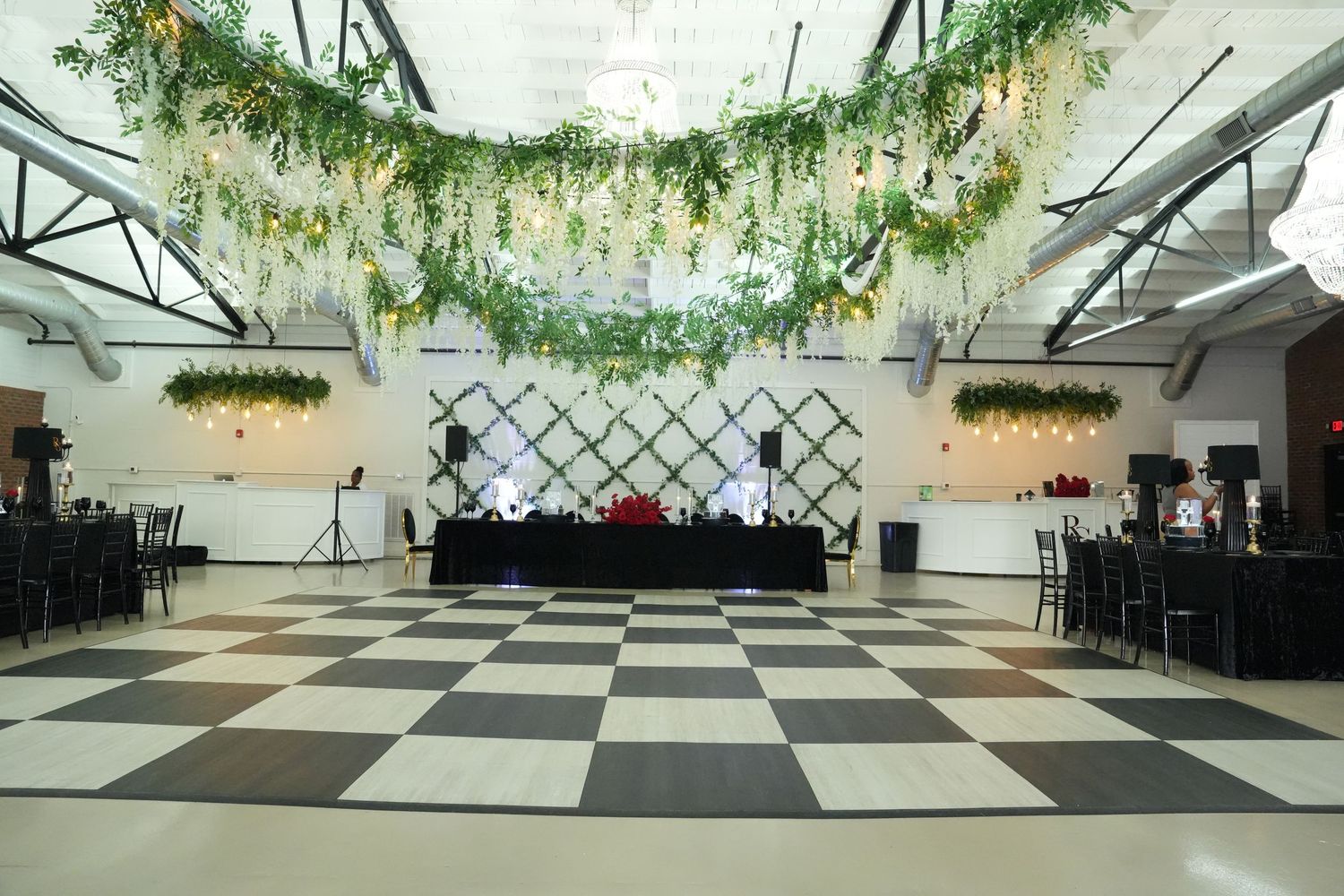 Elegant event venue with checkered dance floor, floral ceiling installation, and decorated DJ booth.
