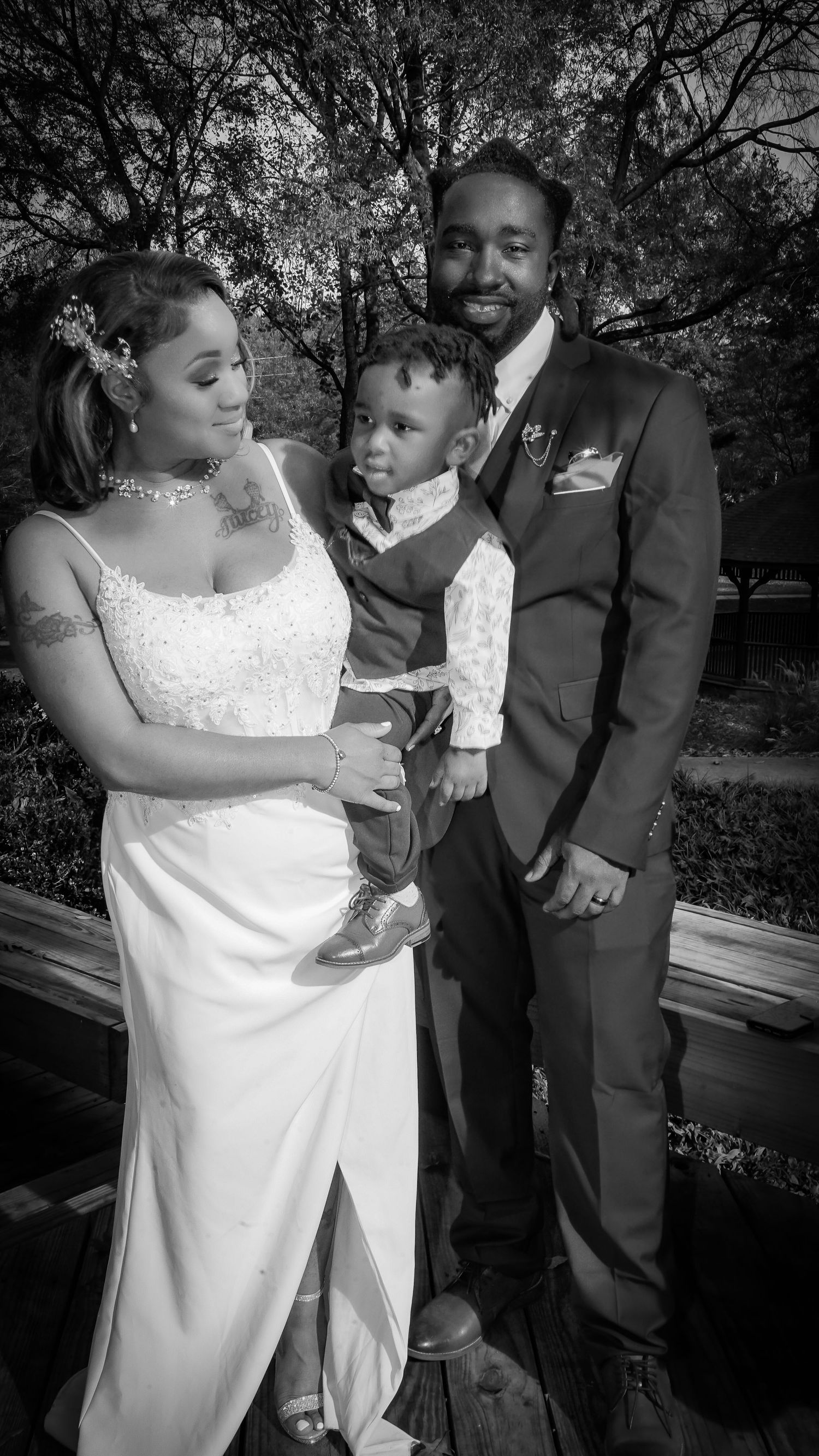 Black and white wedding photo of a bride in white gown and groom in suit holding their young son outdoors.