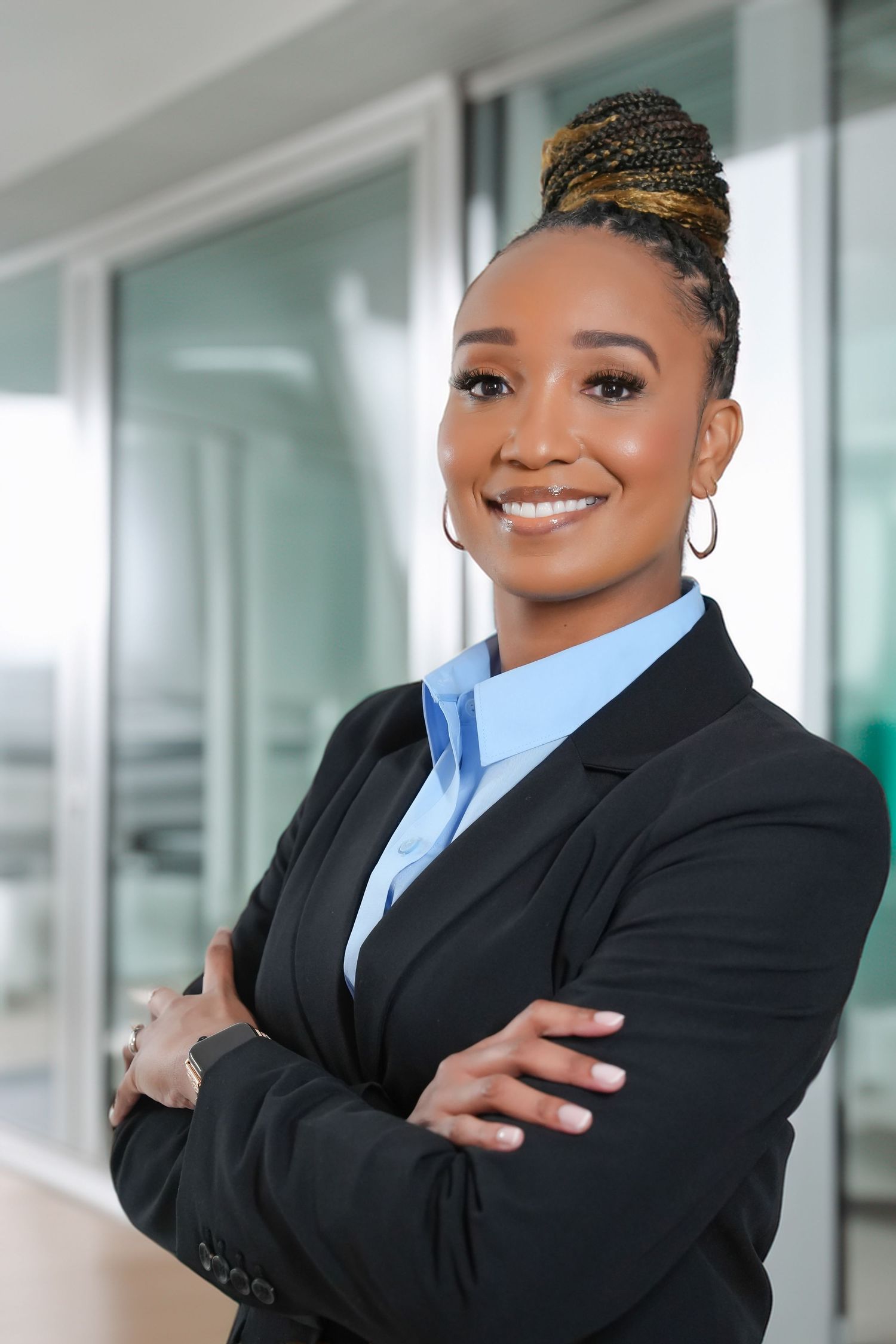 Confident Black businesswoman in black blazer and blue shirt standing with arms crossed smiling in modern office.
