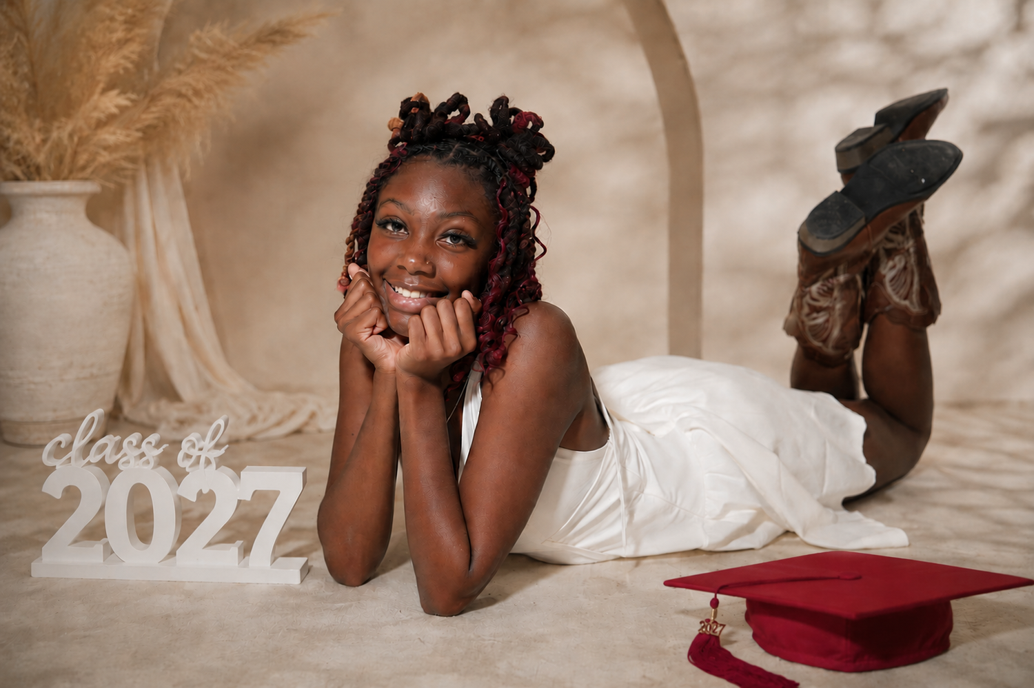 Class of 2027 graduate girl in white dress poses with red cap and cowboy boots in a studio portrait.