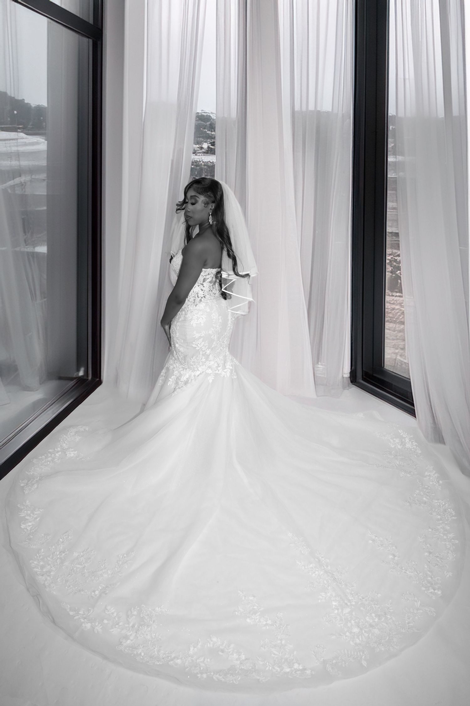Bride in elegant mermaid wedding gown with long lace train and veil stands by large window in black and white photo.