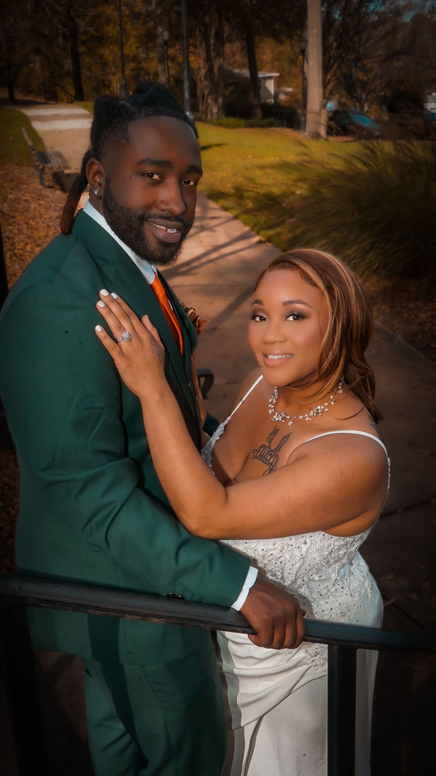 A couple in wedding attire poses by a railing, the groom in a green suit and bride in a white gown.