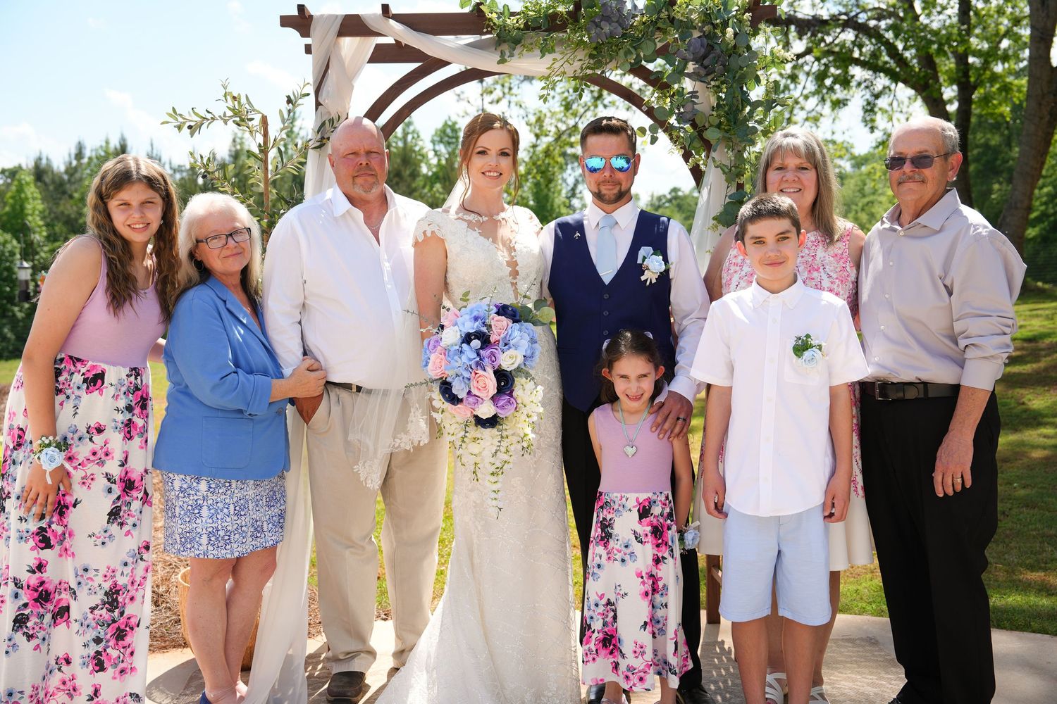 Wedding party poses under floral arch outdoors, bride in white gown with groom in navy suit surrounded by smiling family members.