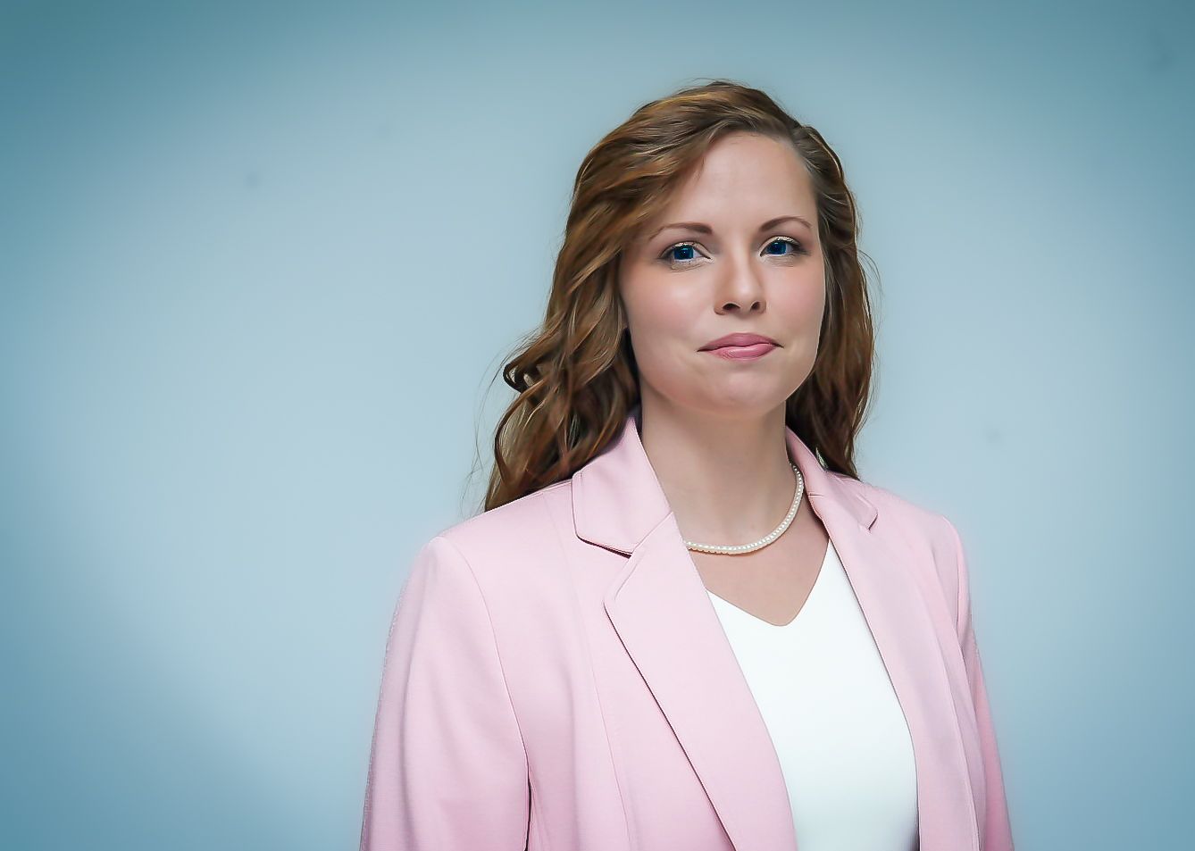 Professional woman in pink blazer and pearl necklace posing against light blue background for business portrait.