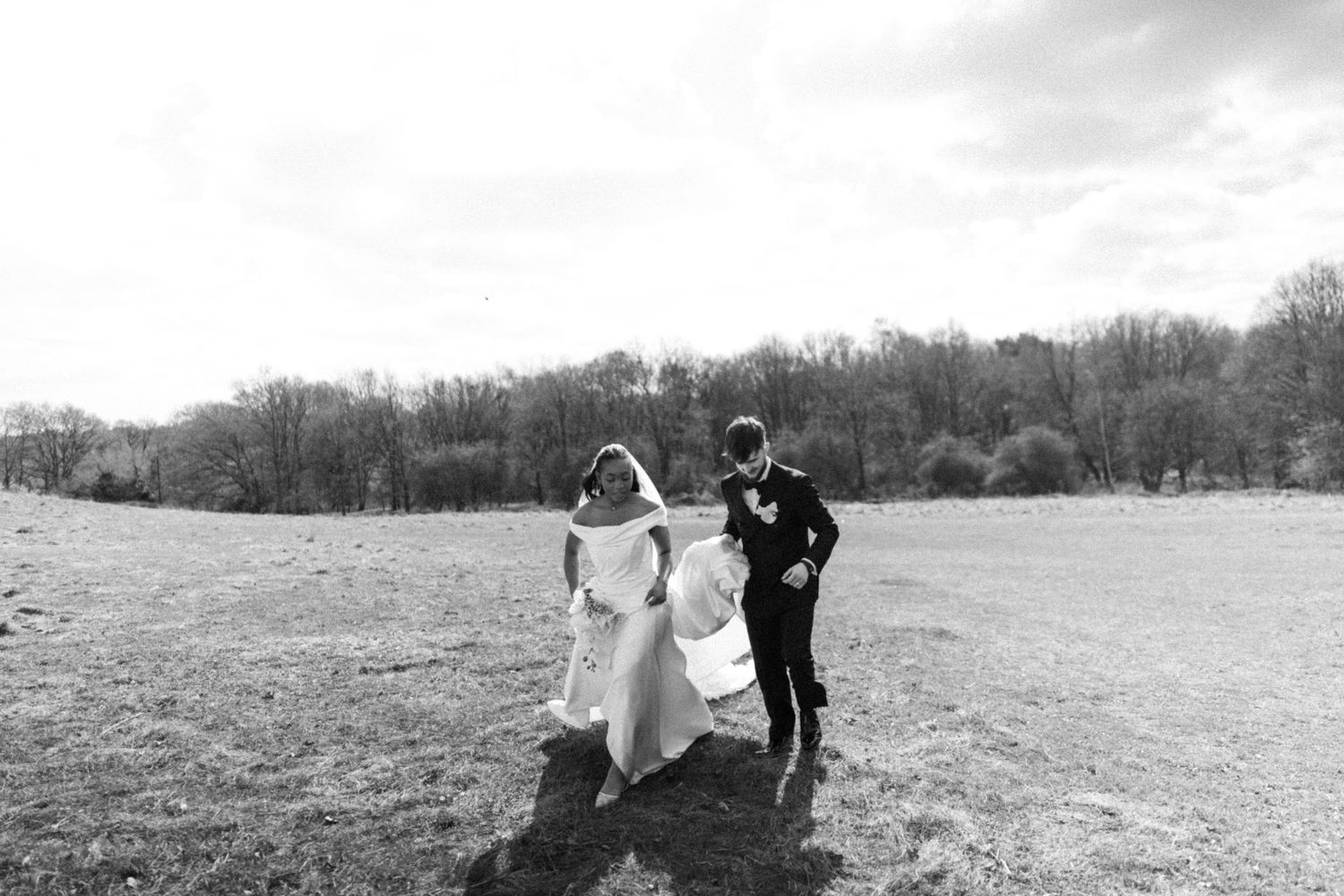 Candid moment (B&W) between bride & groom walking with groom holding the train of the dress