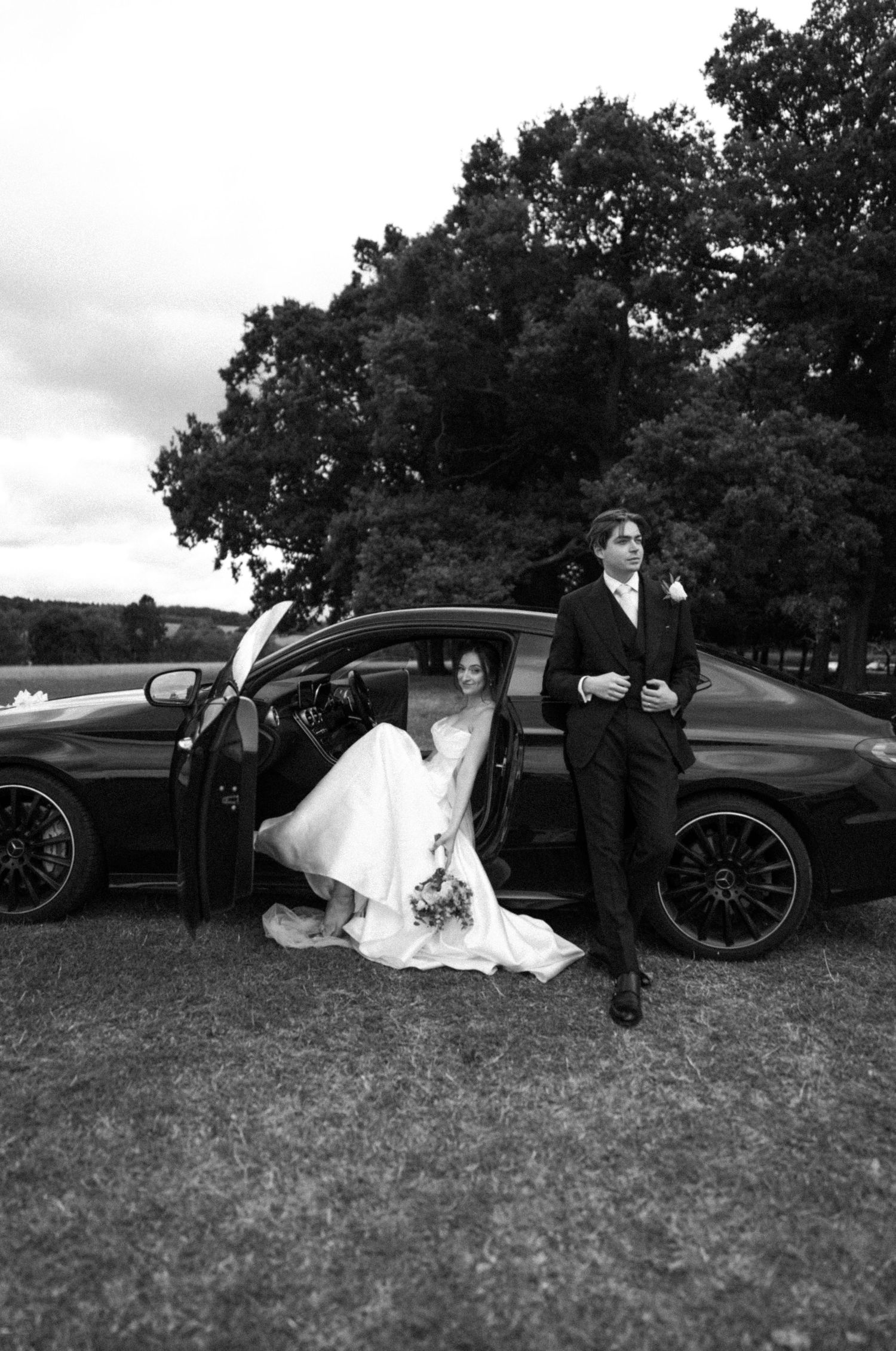 Bride & groom with a wedding car. Bride seated in car and groom leaning against car. Editorial wedding