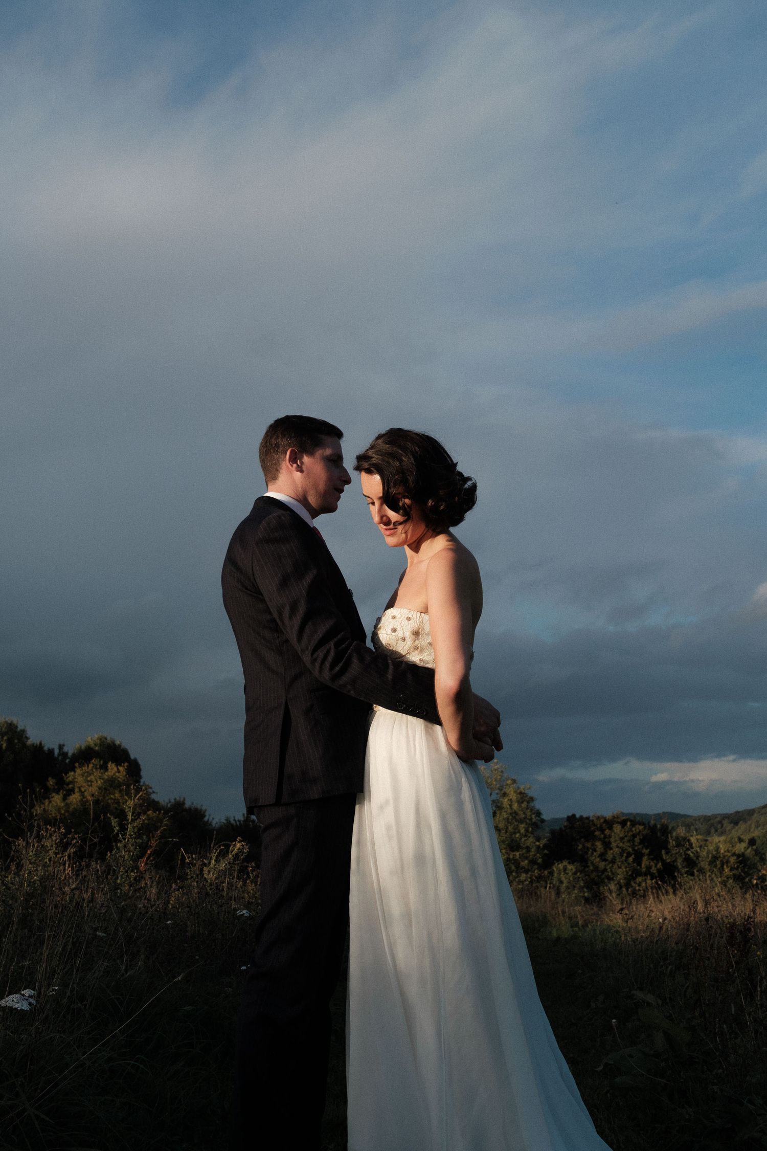 A couple shares an intimate moment at sunset in a field with dramatic cloudy skies and mountains in the background.