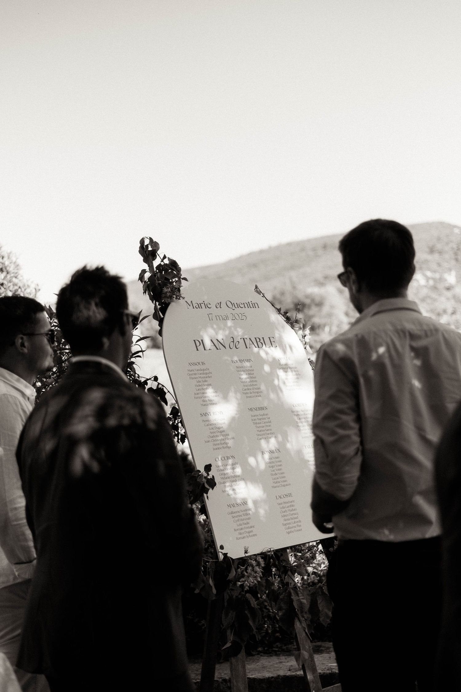 Wedding guests gather around to view the seating chart display at an outdoor venue in black and white photography.