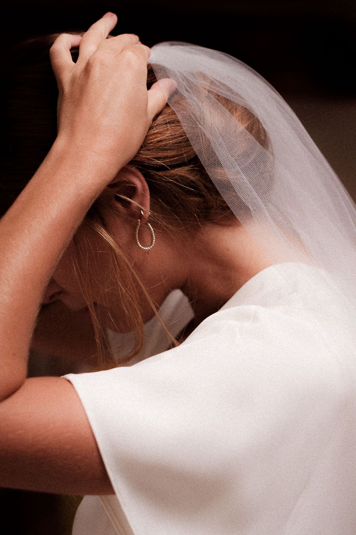 A bride in a white dress adjusts her delicate veil, showing an elegant earring in a candid moment.