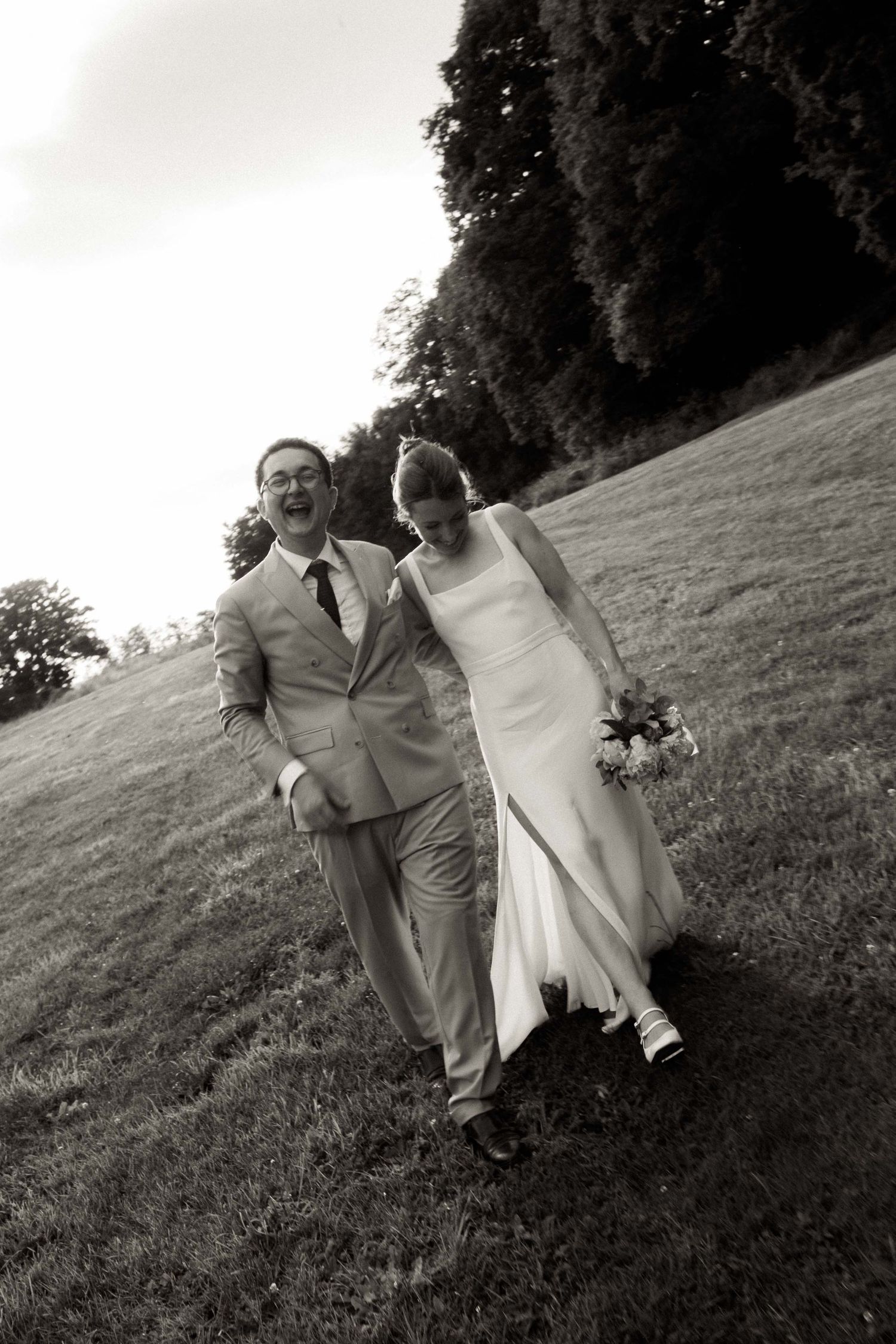 A couple in wedding attire playfully poses on a grassy hillside in black and white photos.