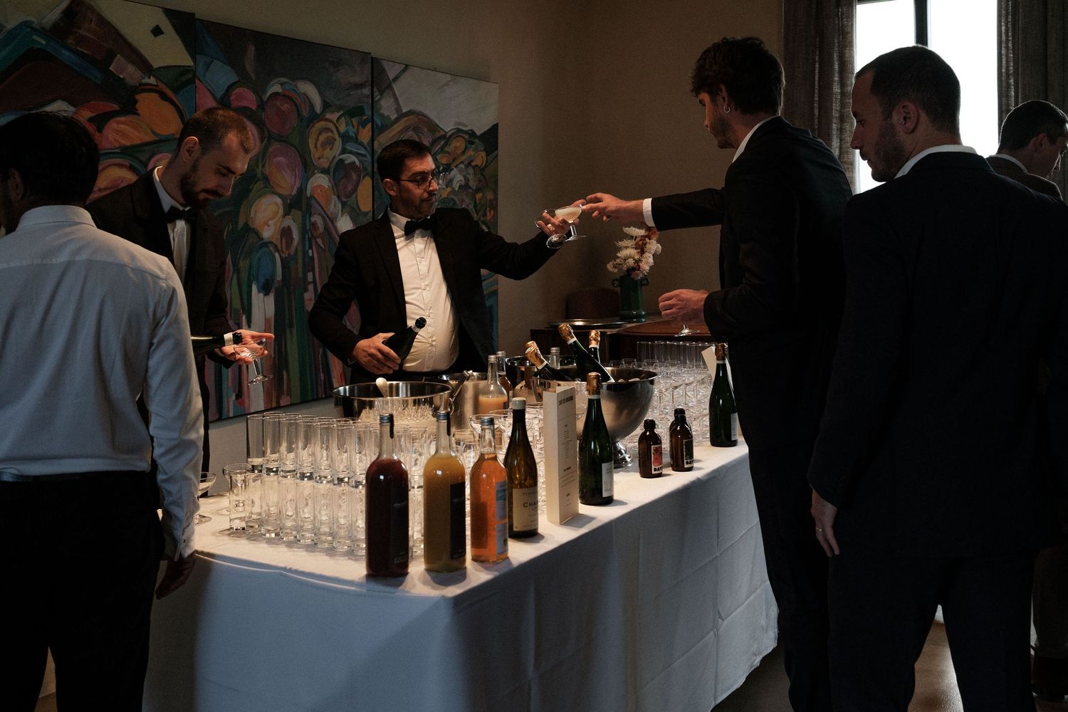 Wine bottles and glasses line a long table at a tasting event with attendees examining selections in dim lighting.