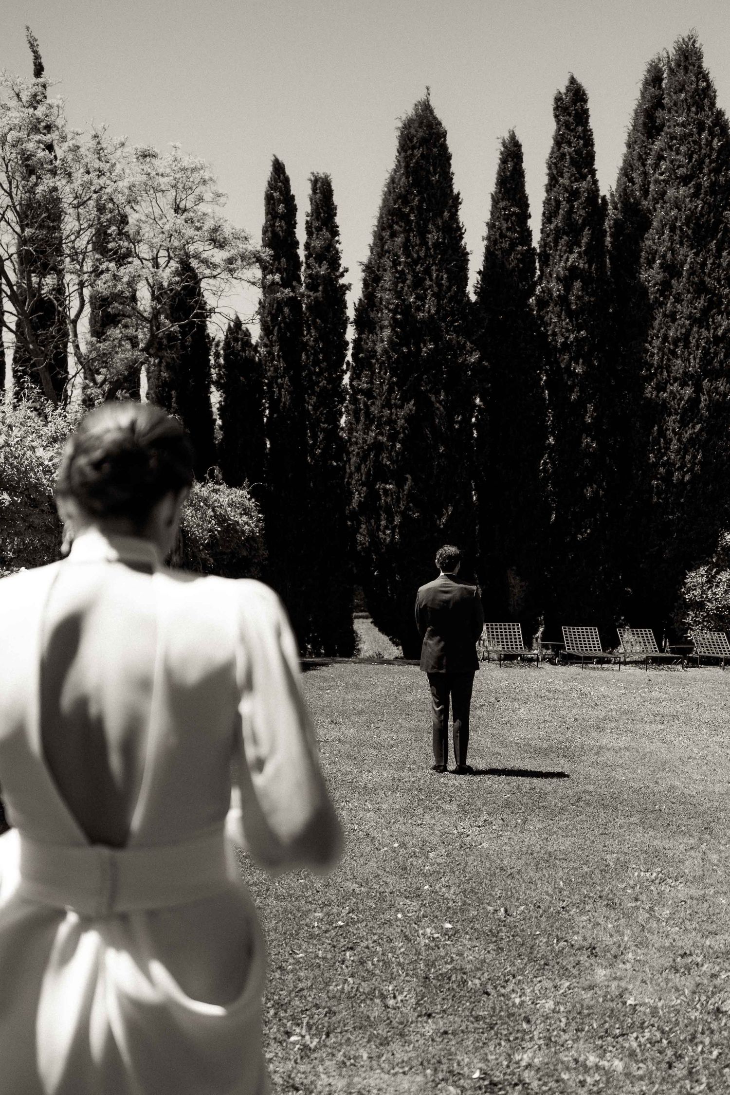 A black and white wedding photo showing a dramatic first look scene with tall cypress trees.