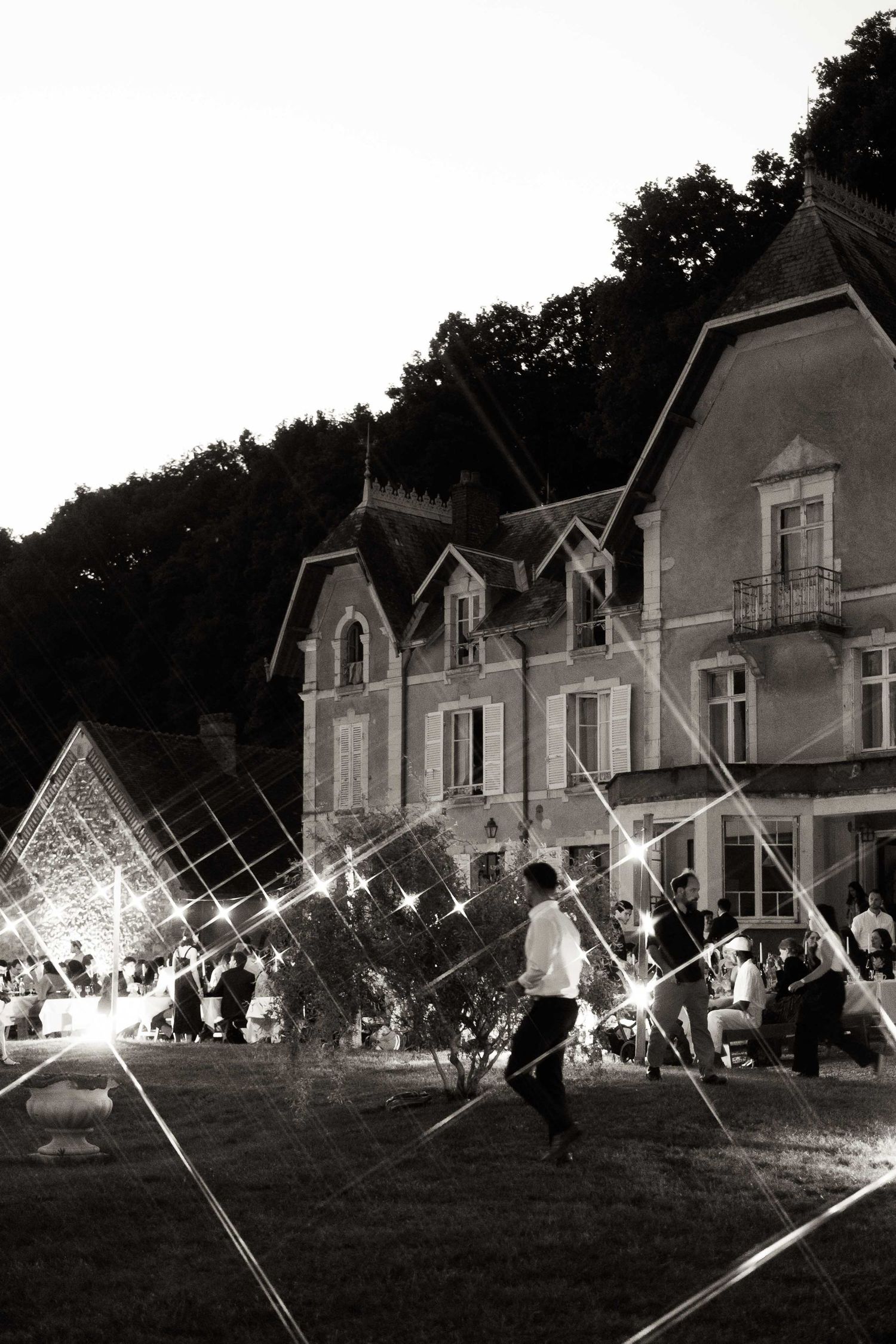 A black and white panoramic sequence showing teepees and people gathering on a lawn in front of historic houses at night.