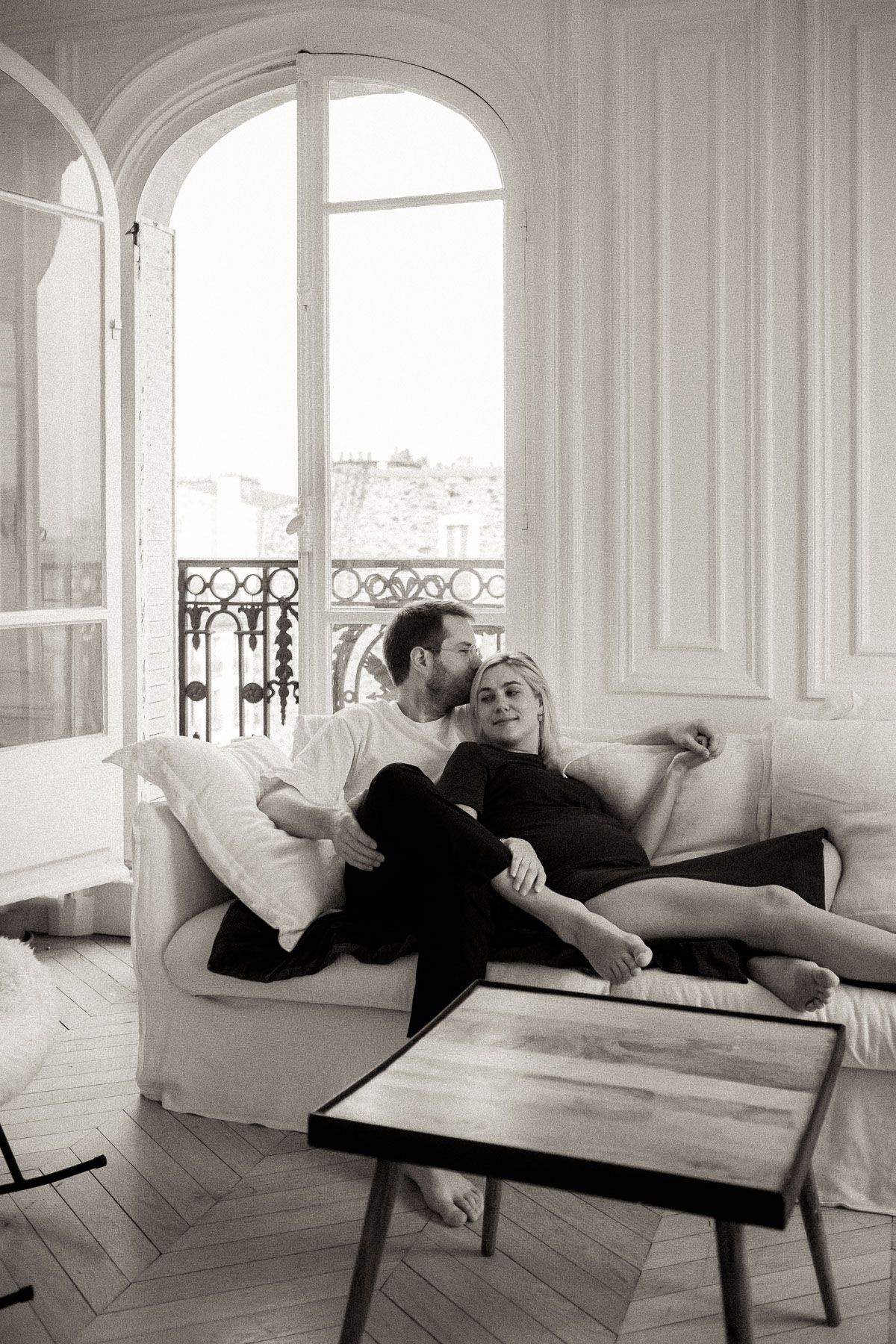 A black and white photo series shows someone reclining on a sofa in an elegant room with large windows and herringbone floors.