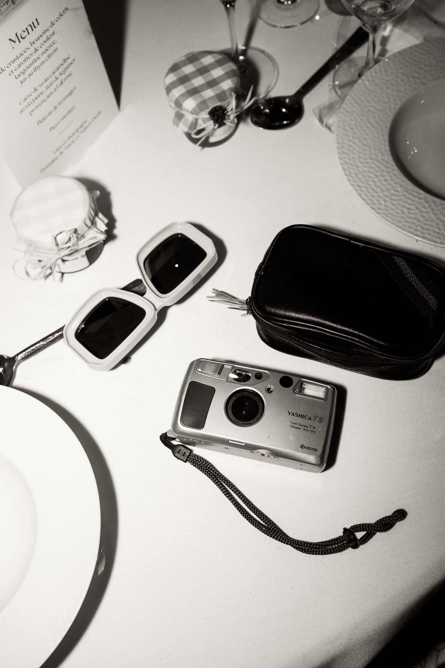 A black and white photo showing a compact camera, sunglasses, and table settings in an artistic composition.