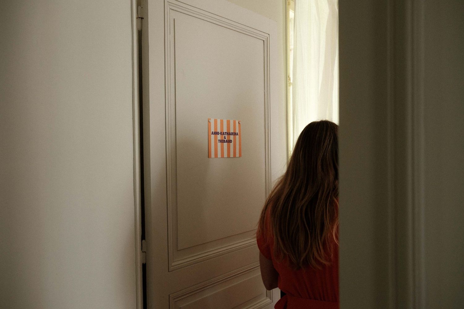A person in a red sweater stands in a dimly lit hallway facing a white door with a small orange sign.