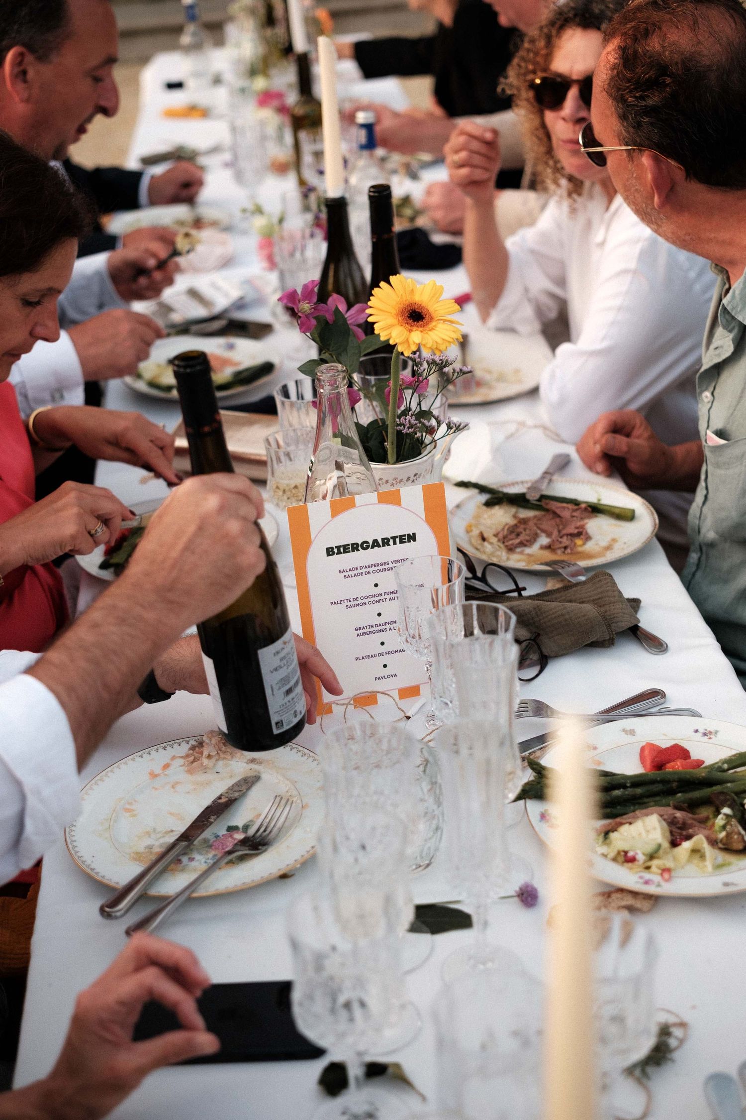 Diners seated at a long table with wine glasses and plates enjoying a meal at what appears to be a dinner party.