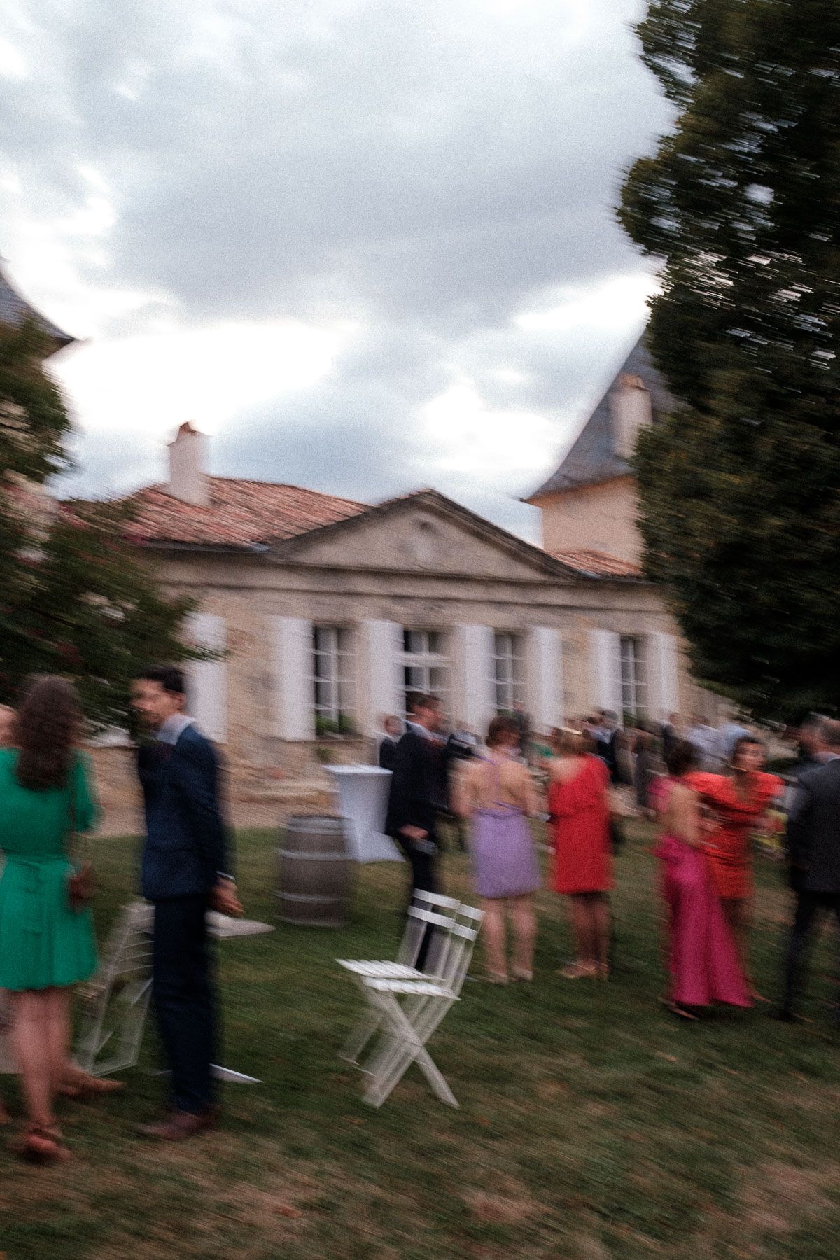 A line of wedding guests in formal attire stand on a grassy hill outside a stone building during an outdoor celebration.
