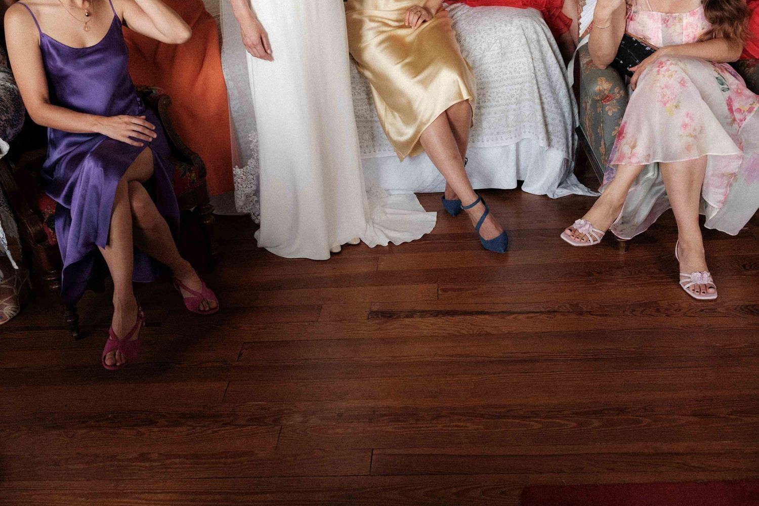 A group of people in formal dresses and gowns sitting with their feet visible on a dark hardwood floor.